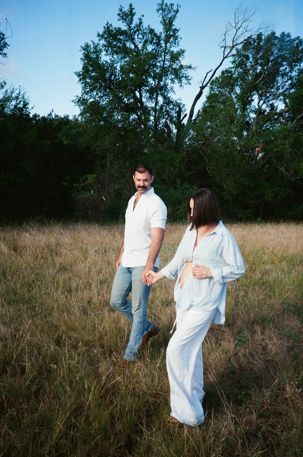 A couple poses in a field at dusk wearing white and denim outfits.