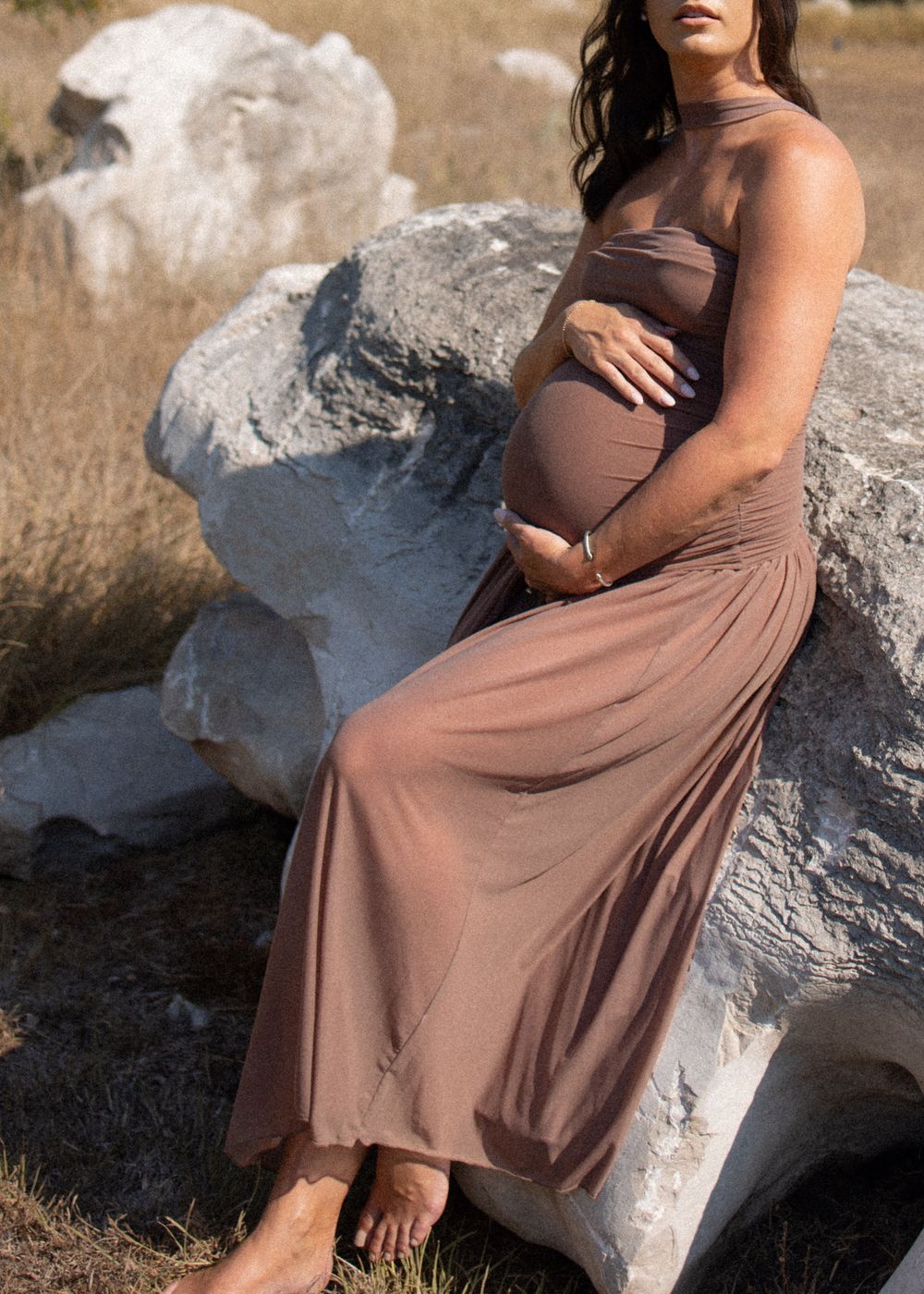 Maternity photo of pregnant woman in brown dress sitting on large rock.