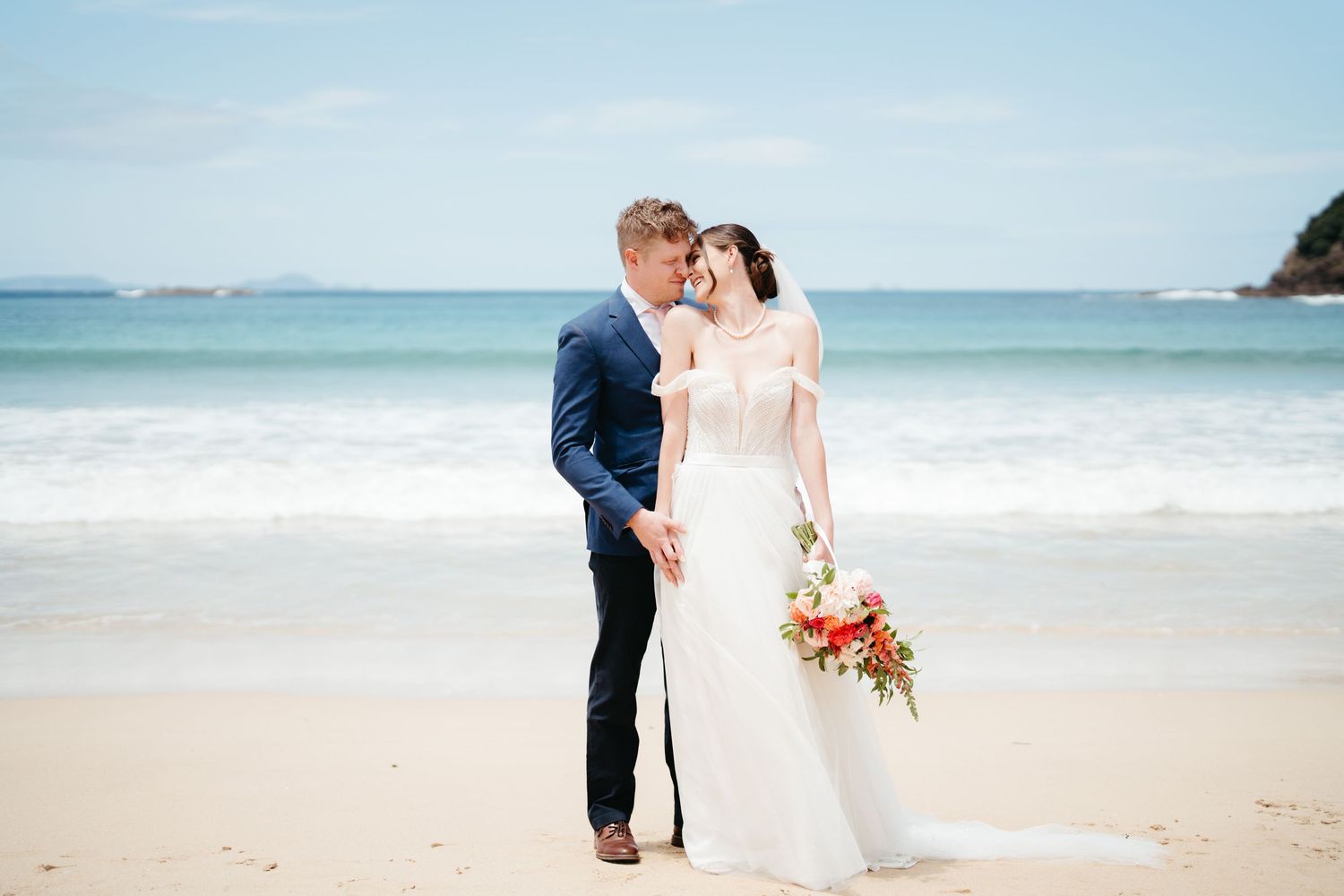 A couple shares a romantic moment on a pristine beach in wedding attire with coral bouquet.