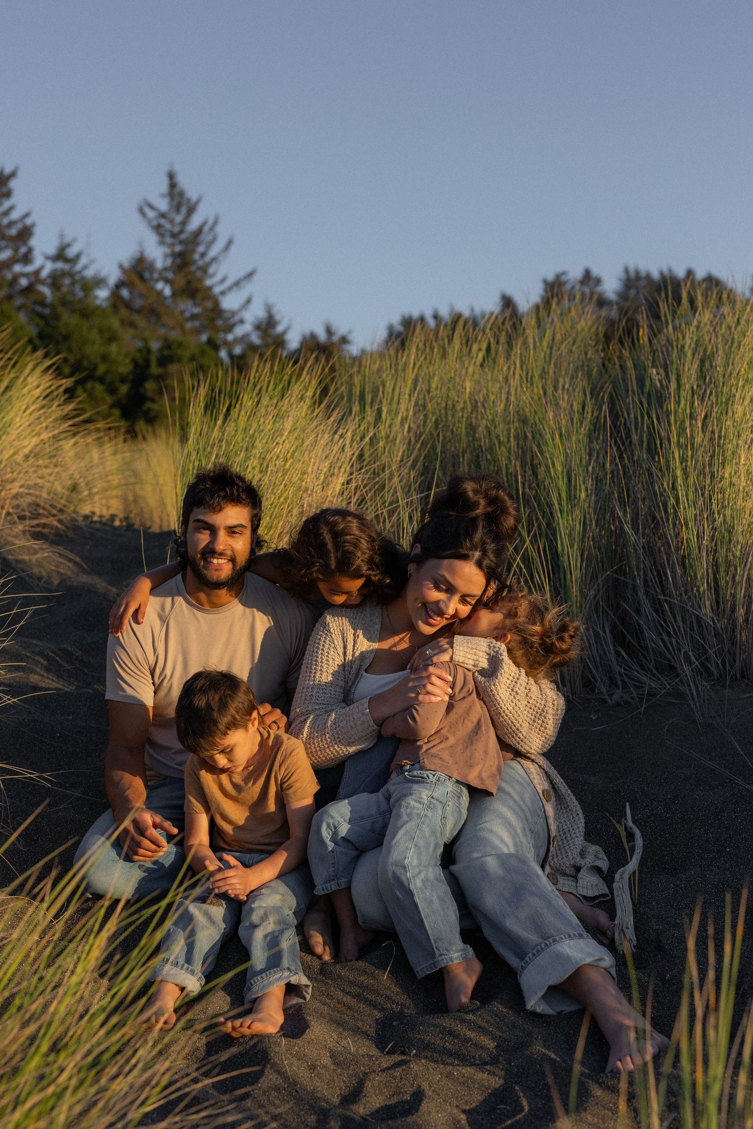 A family sits together among tall beach grass on the Oregon Coast during golden hour at sunset while sharing joyful moments together.