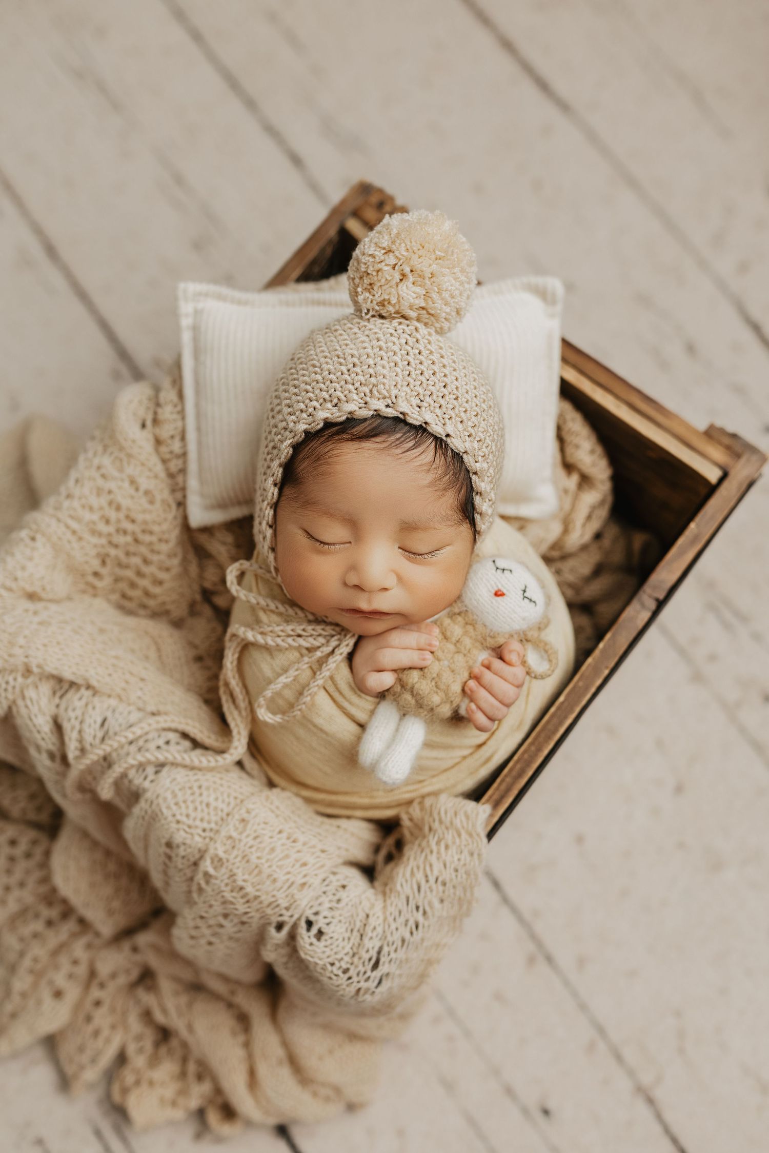 Newborn in beige knit hat sleeping peacefully in wooden box while holding small plush toy.