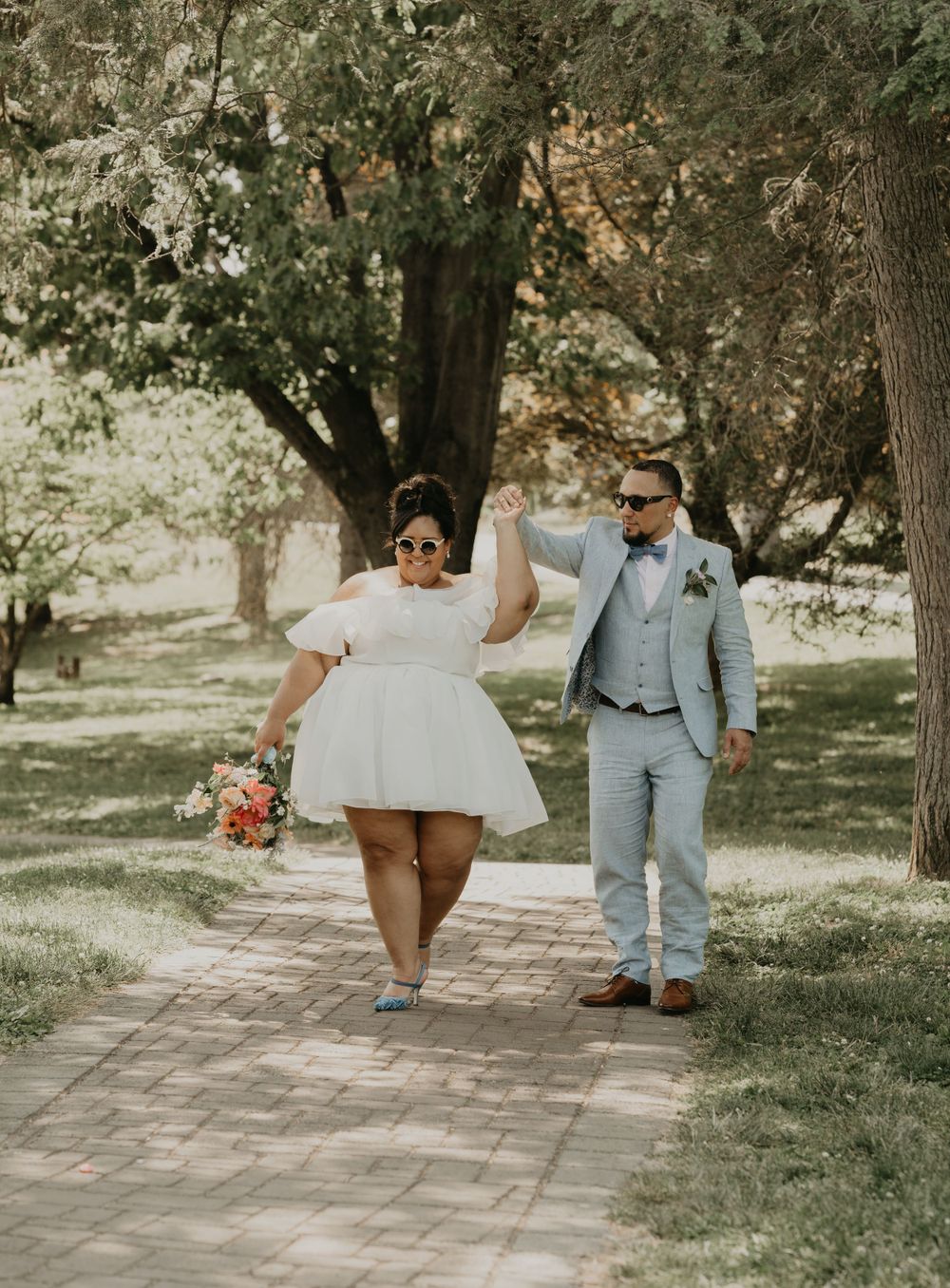 Newlyweds walking along a tree-lined brick pathway in a park.