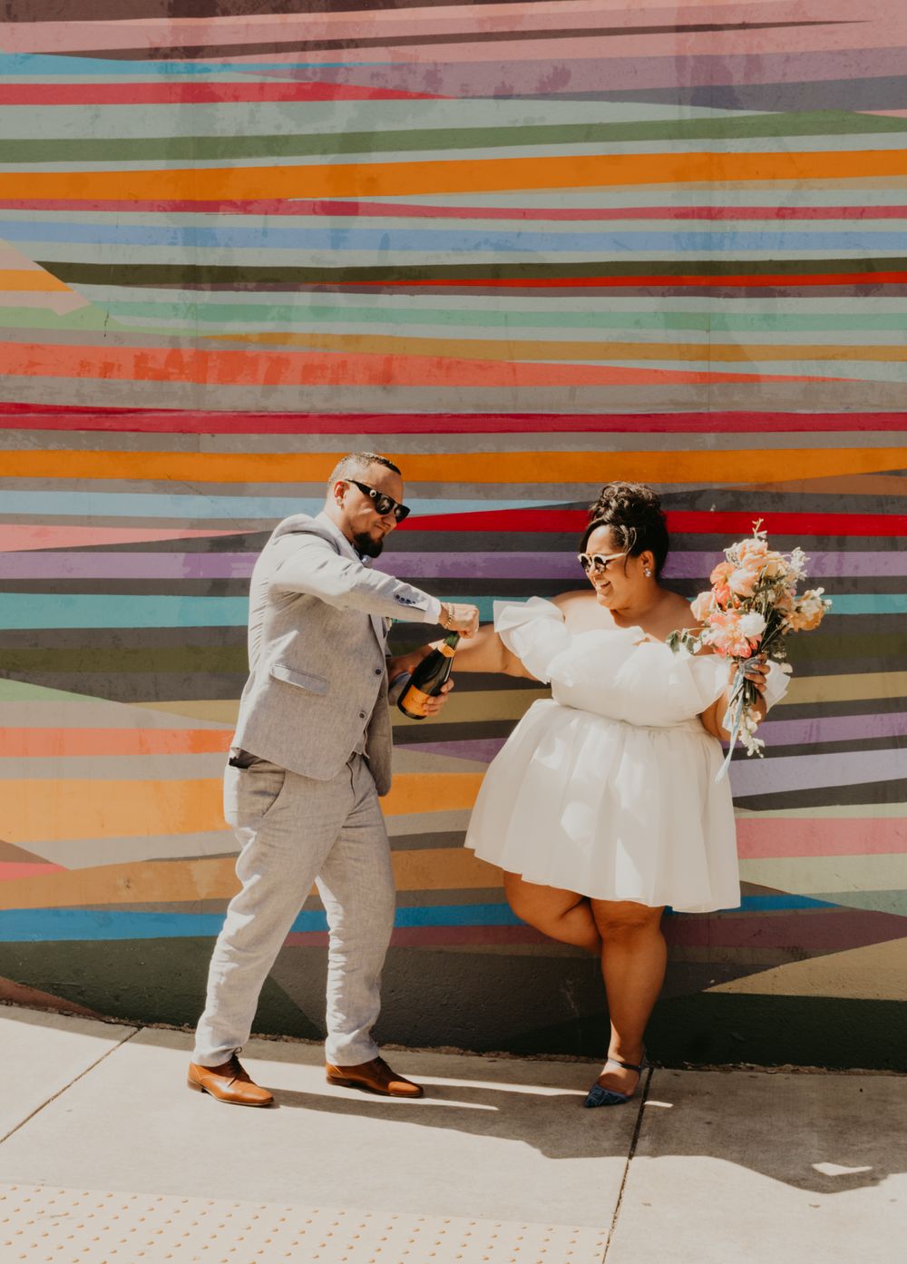 Newlyweds strike playful dance poses against a vibrant multicolored wall.