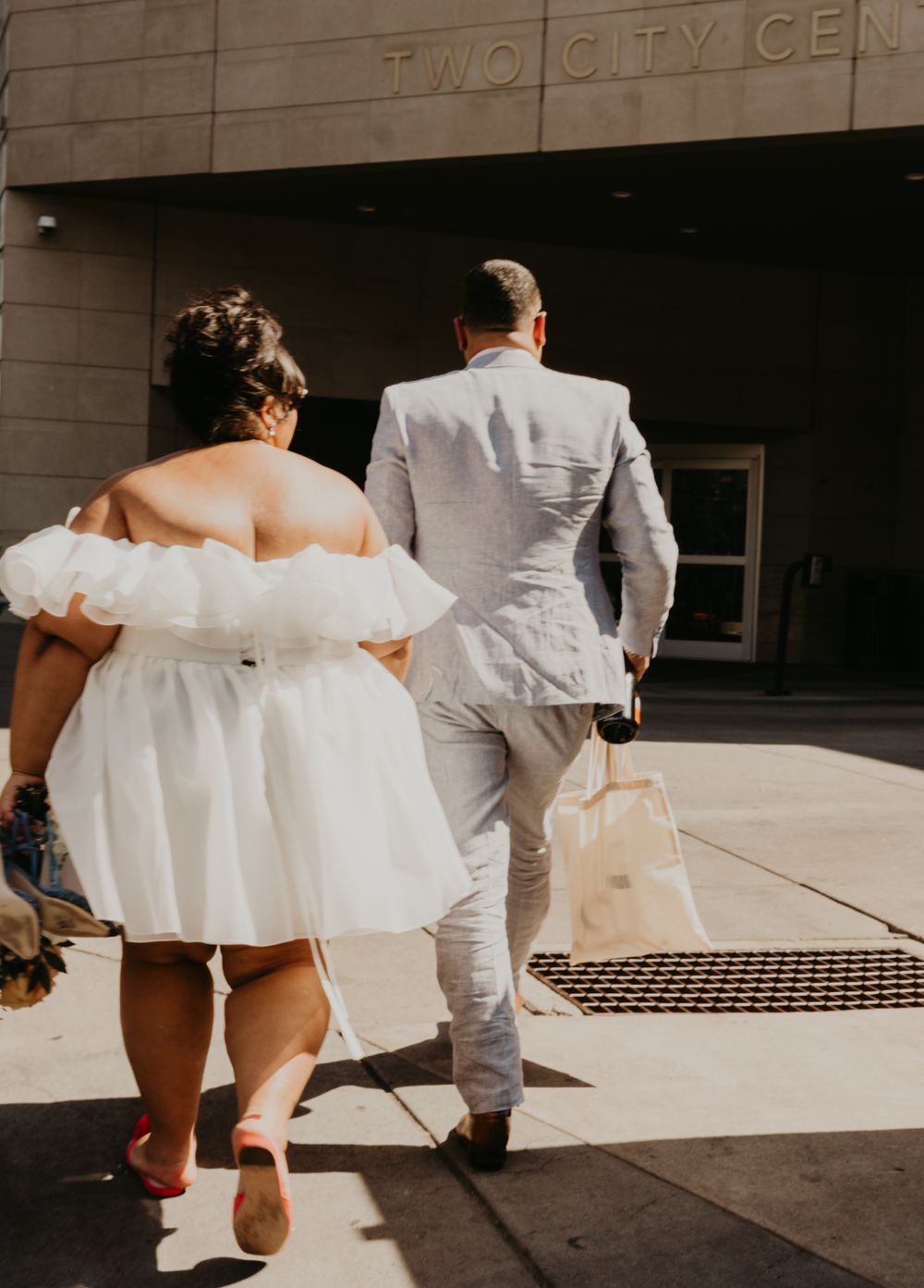 A couple in wedding attire walking towards a city center entrance in bright sunlight.