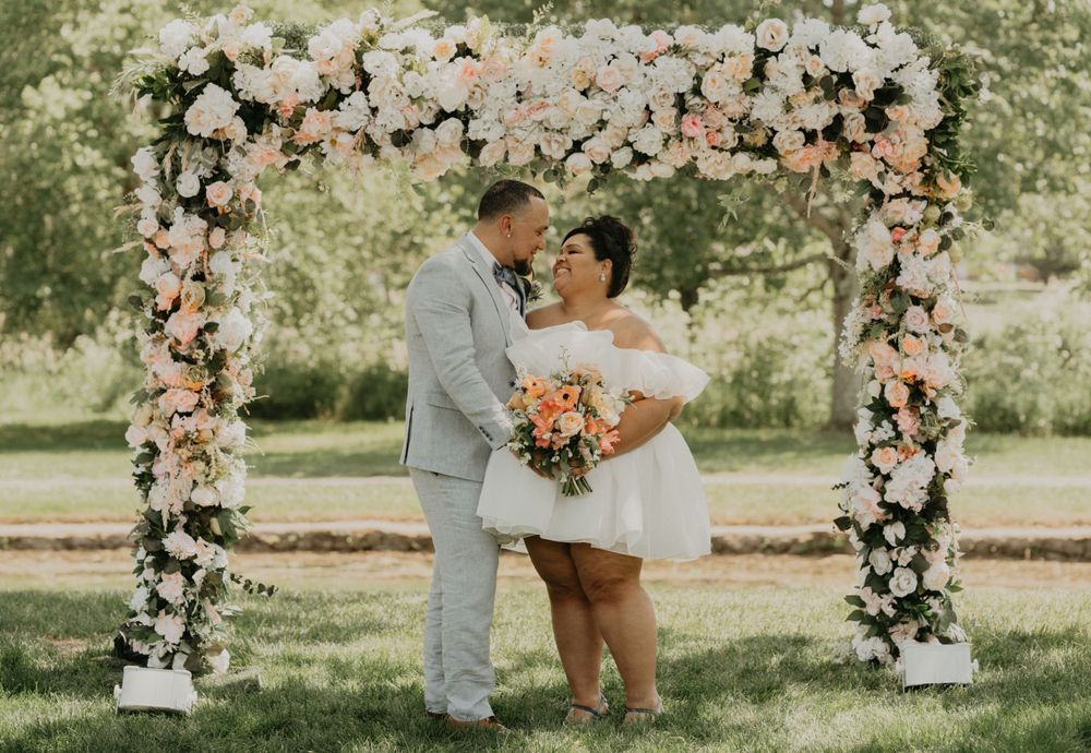 A couple embraces under a floral arch adorned with pink and white flowers.