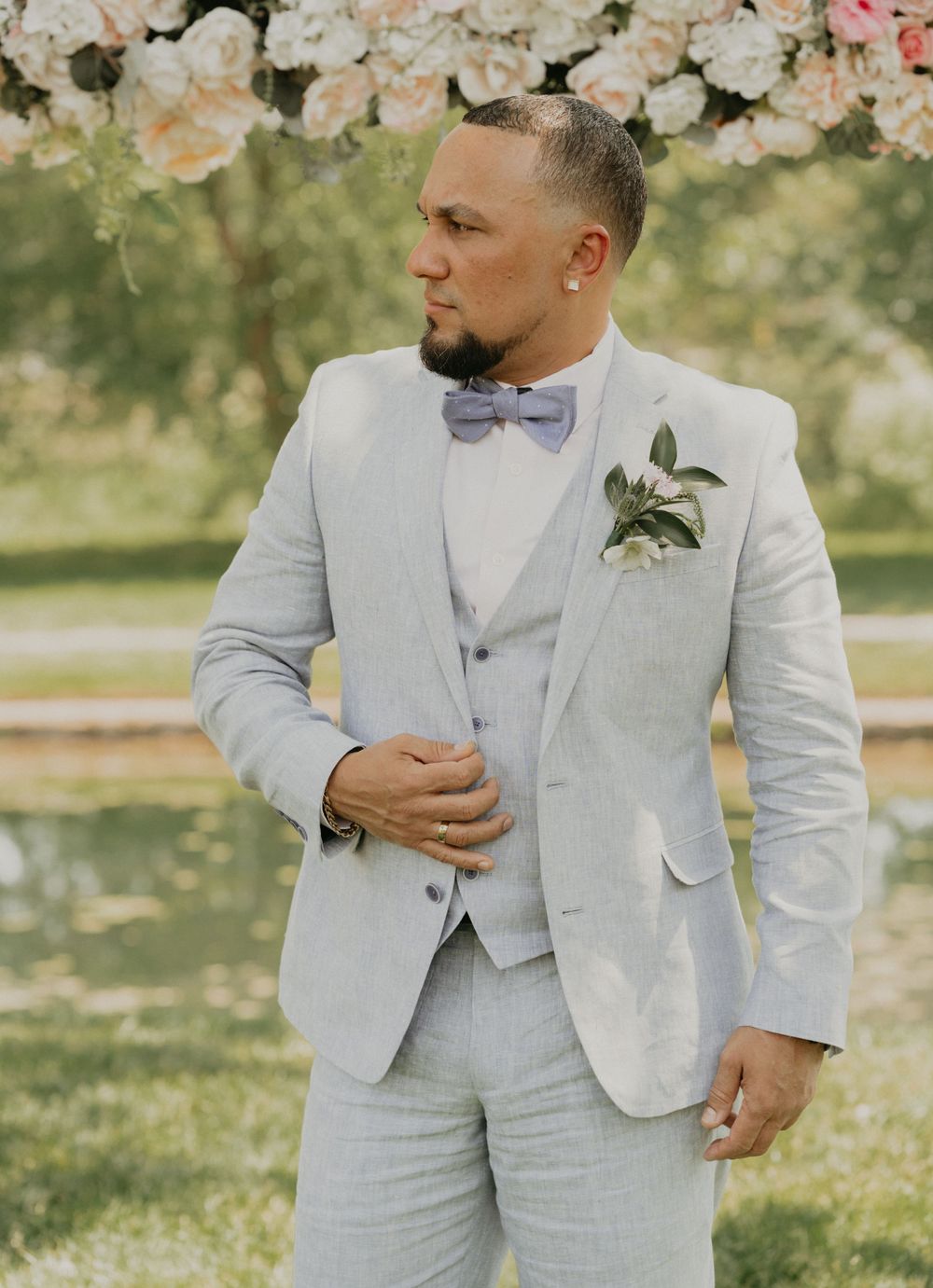 A person in a light grey suit with bow tie stands in a garden setting.