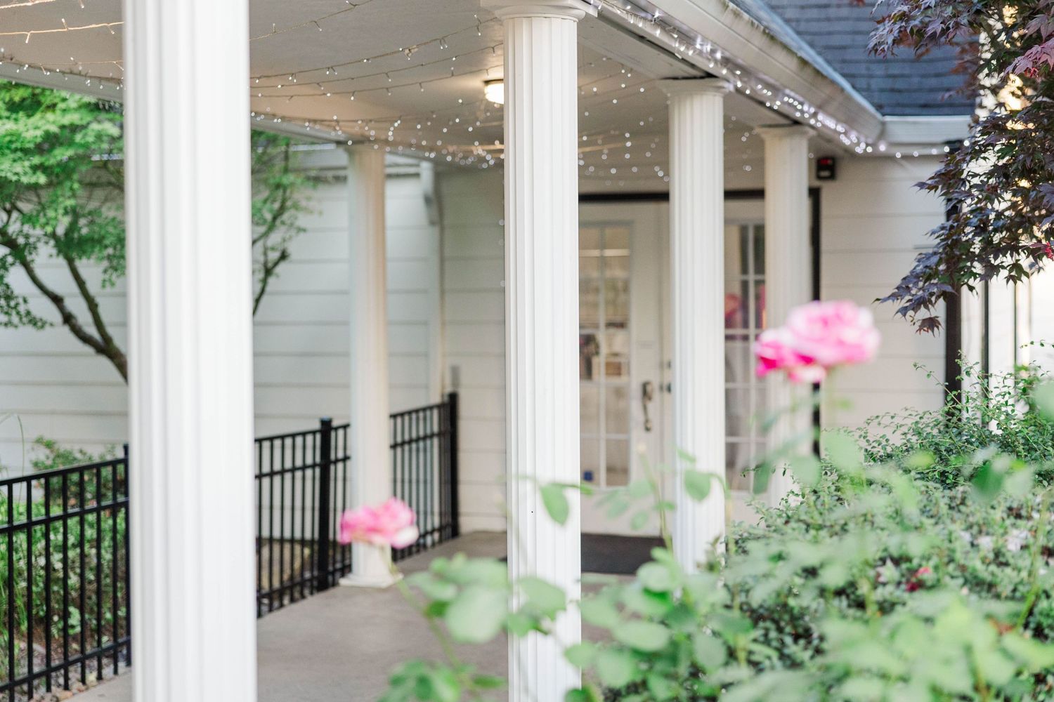 Elegant white colonial porch with tall columns, string lights, and pink roses in lush garden setting.
