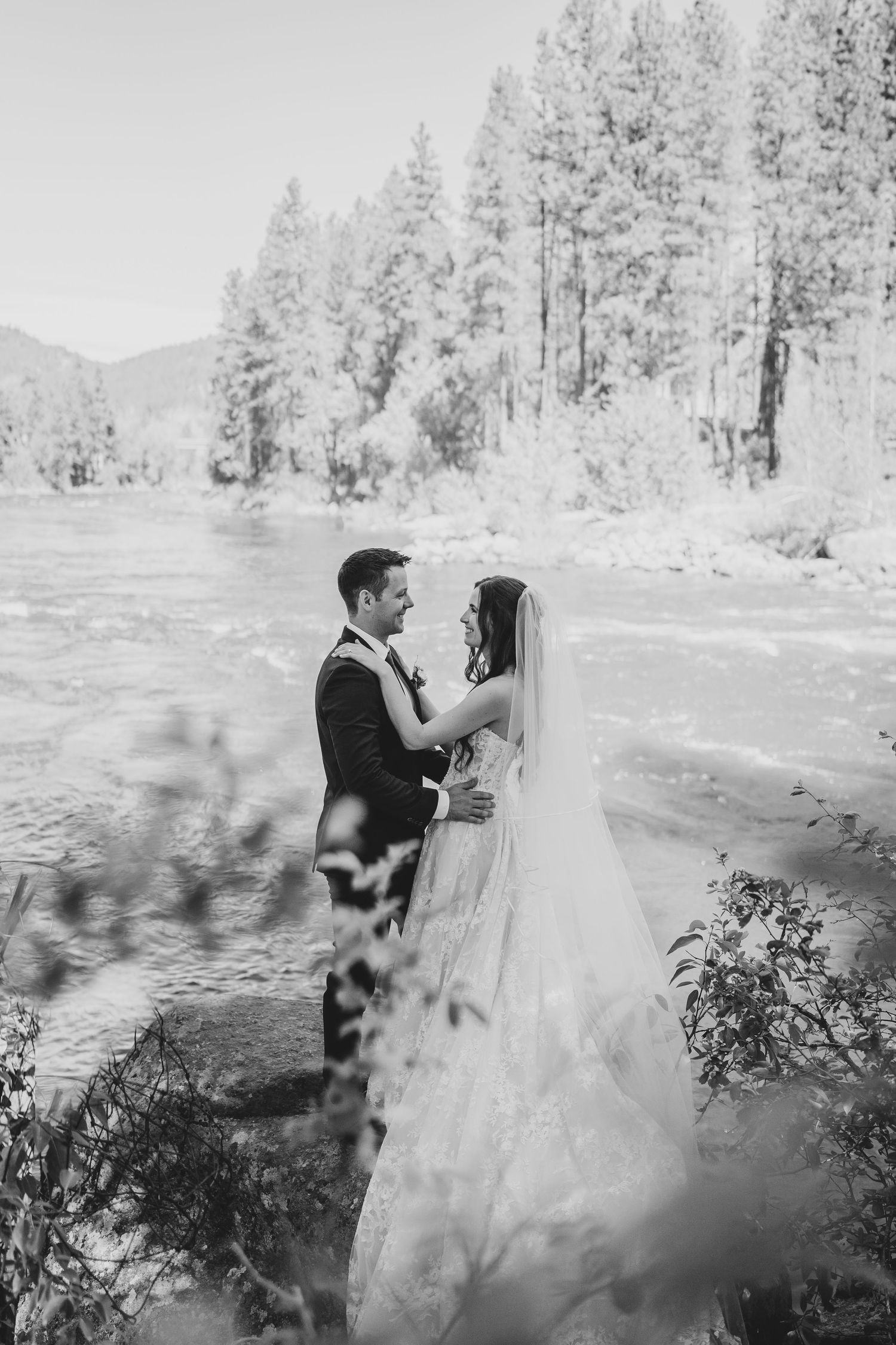 Couple in wedding attire standing by scenic river with pine trees in black and white.