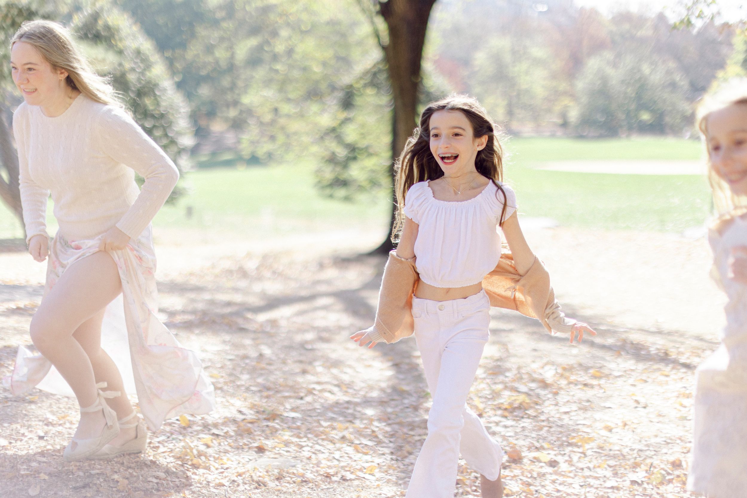 Group in coordinated white and cream outfits runs joyfully through a sunlit park.