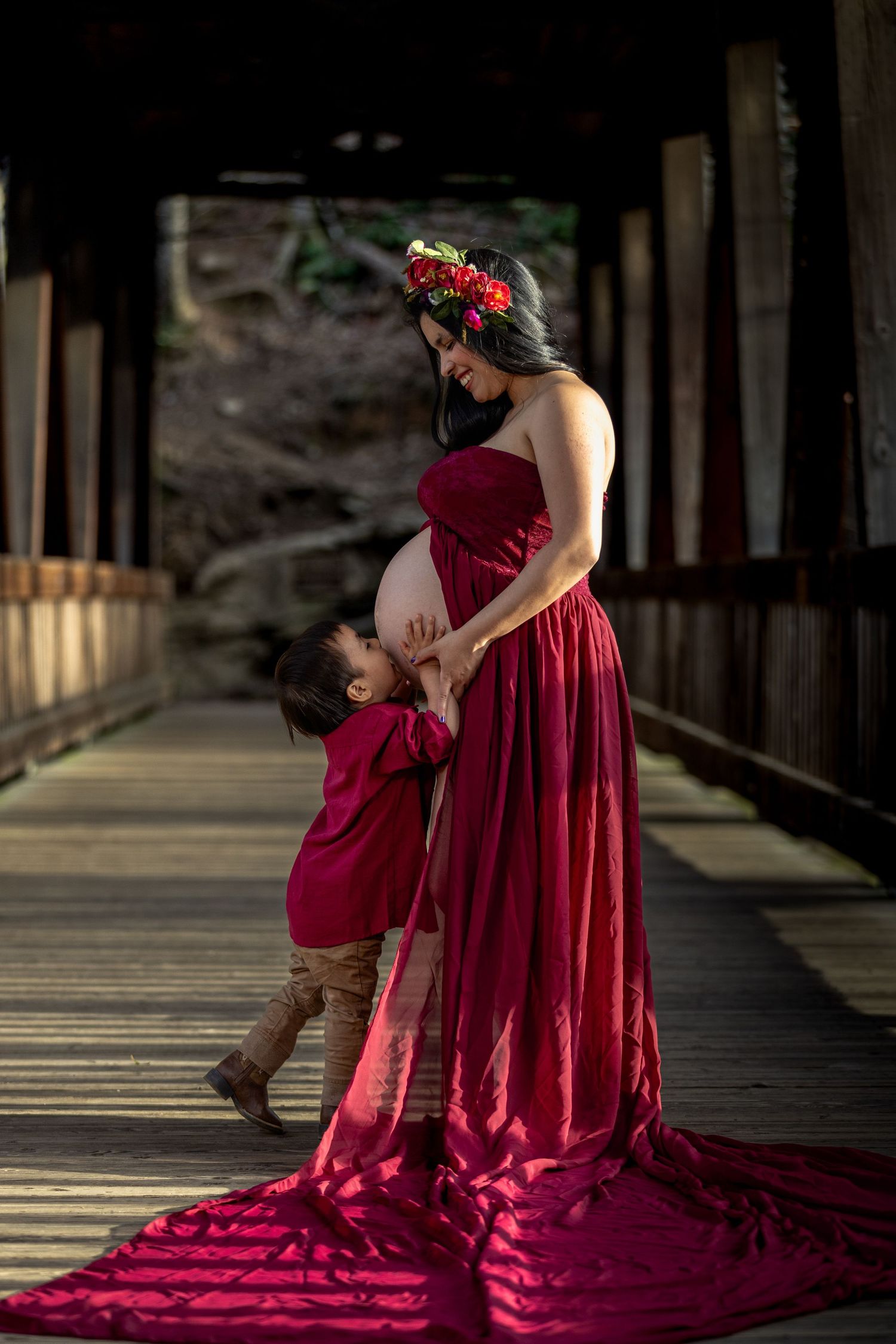 Dramatic maternity photos in flowing red dress on wooden bridge with natural lighting.