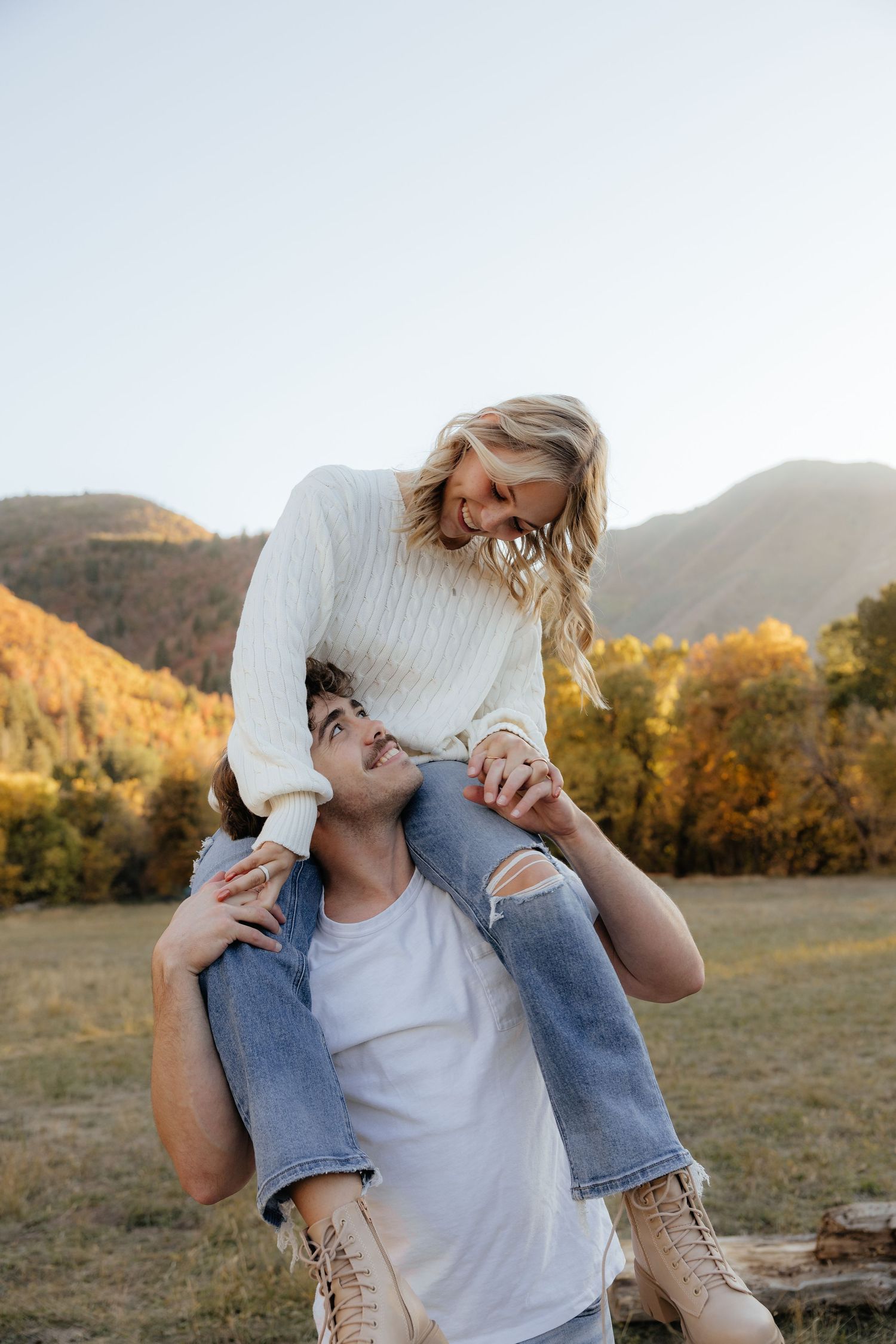 Couple laughing outdoors, woman sitting on man's shoulders in autumn mountain landscape.