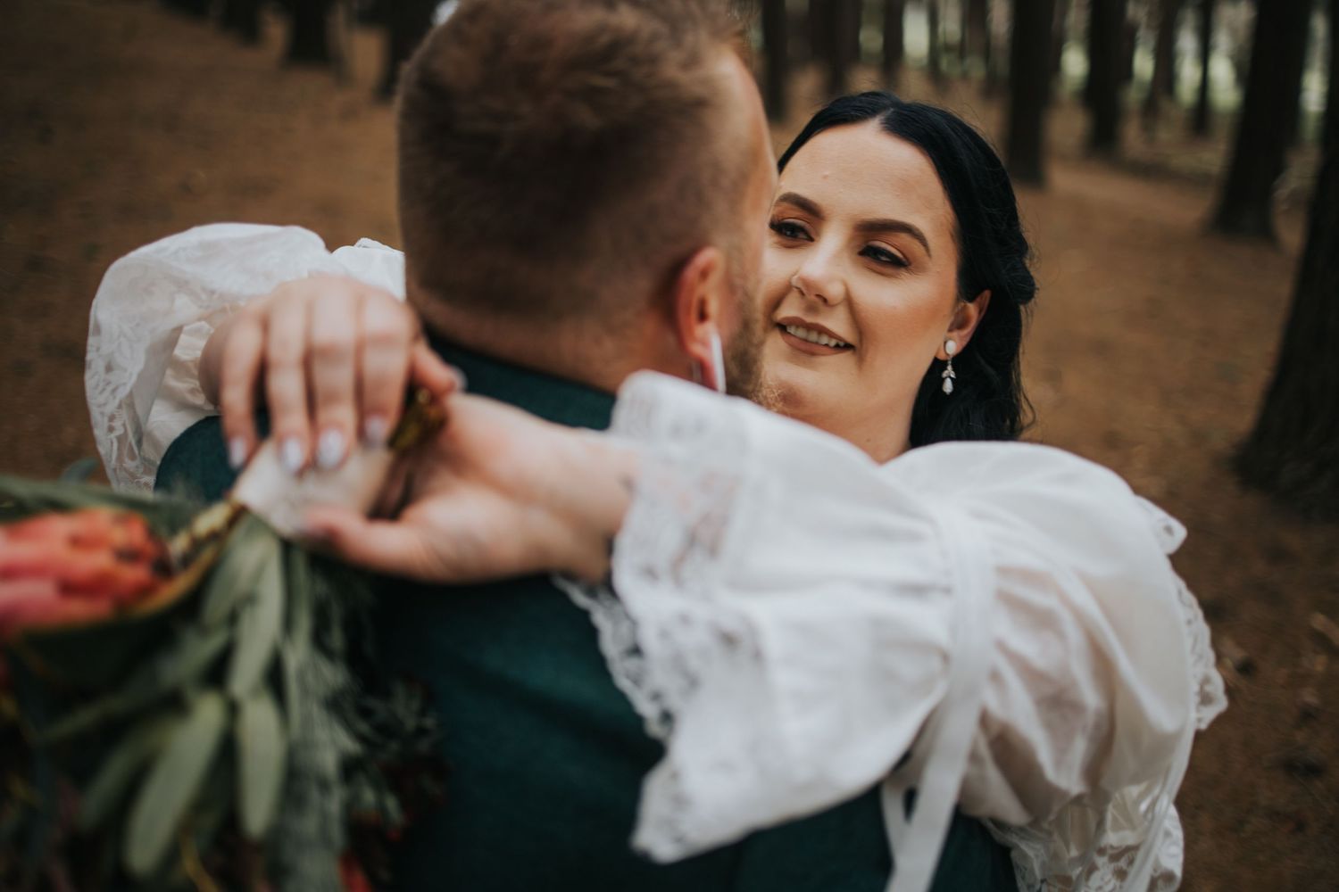 Smiling dark-haired bride embraces groom in pine forest holding colorful native flower bouquet.
