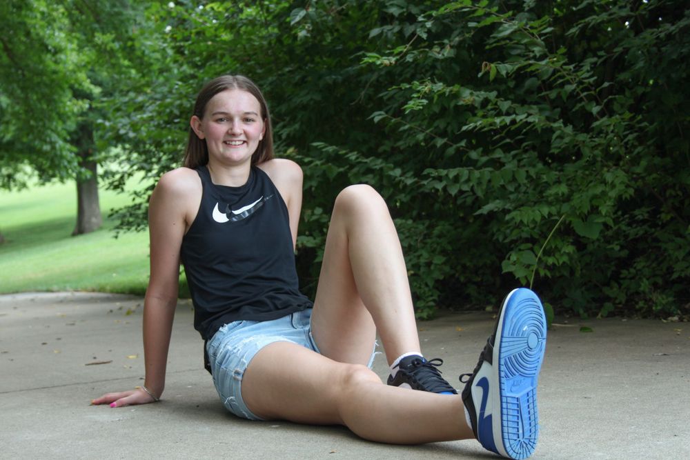 Girl smiling sitting on the ground in front of trees on a trail.