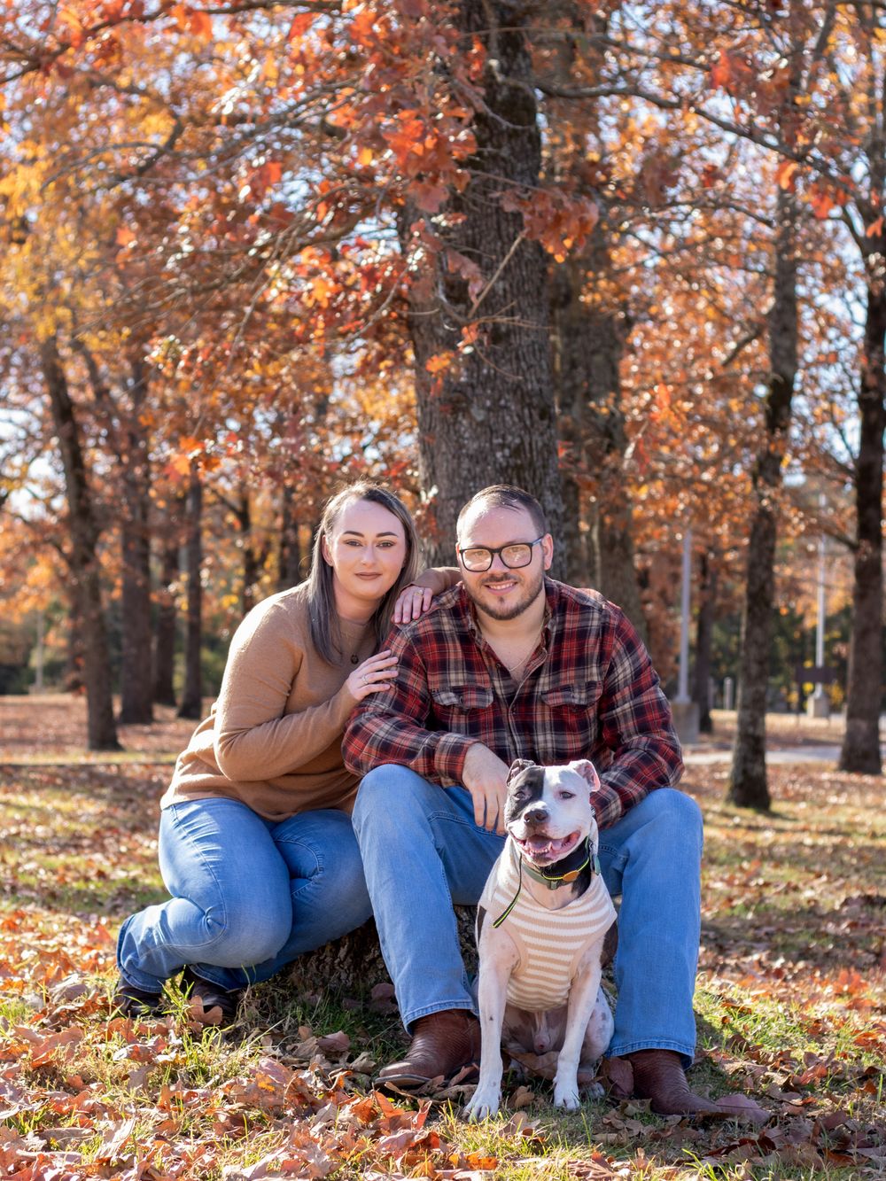 Husband and while smiling with dog in the fall leaves.
