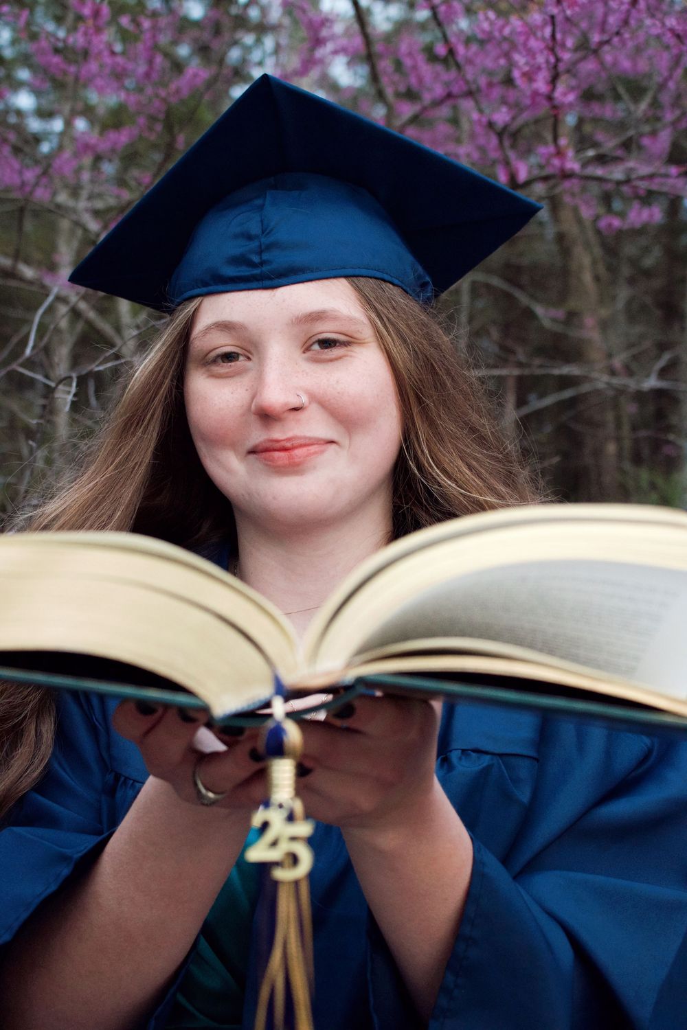 Girl wearing cap and gown holding book in front of purple trees for her senior pictures.