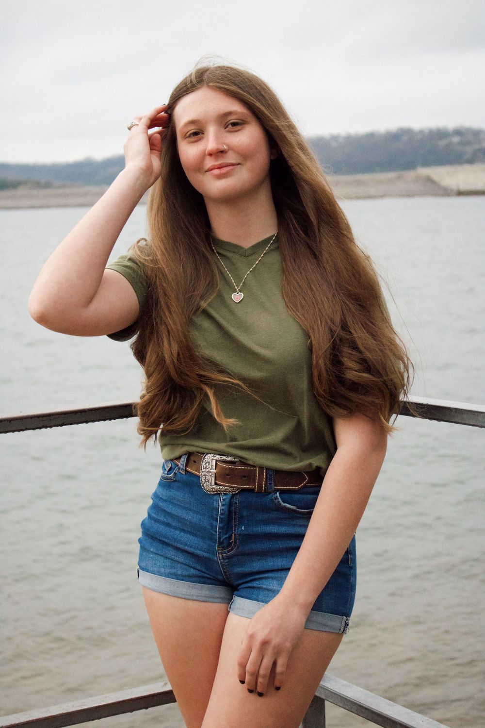 Girl pushing her hair out of her face while smiling at the camera in front of the lake for her senior pictures.