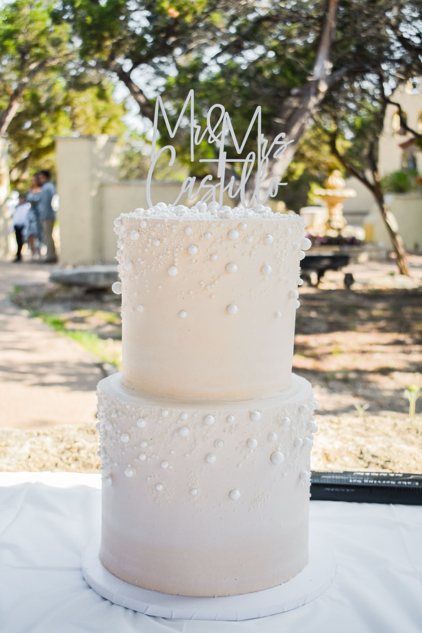 Two-tier white wedding cake with pearl details and a 'Mr & Mrs' topper displayed outdoors.