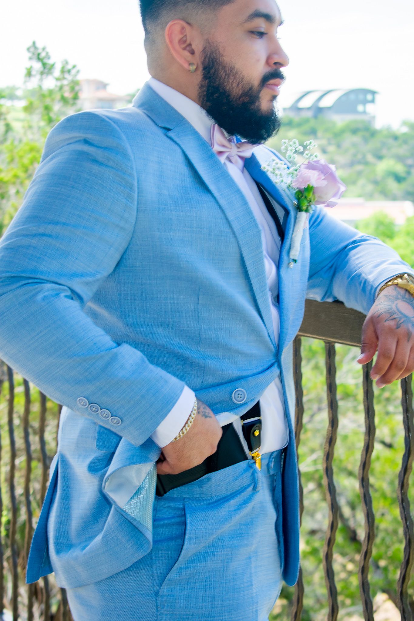 A person in a light blue suit with a pink boutonniere stands against an outdoor railing.