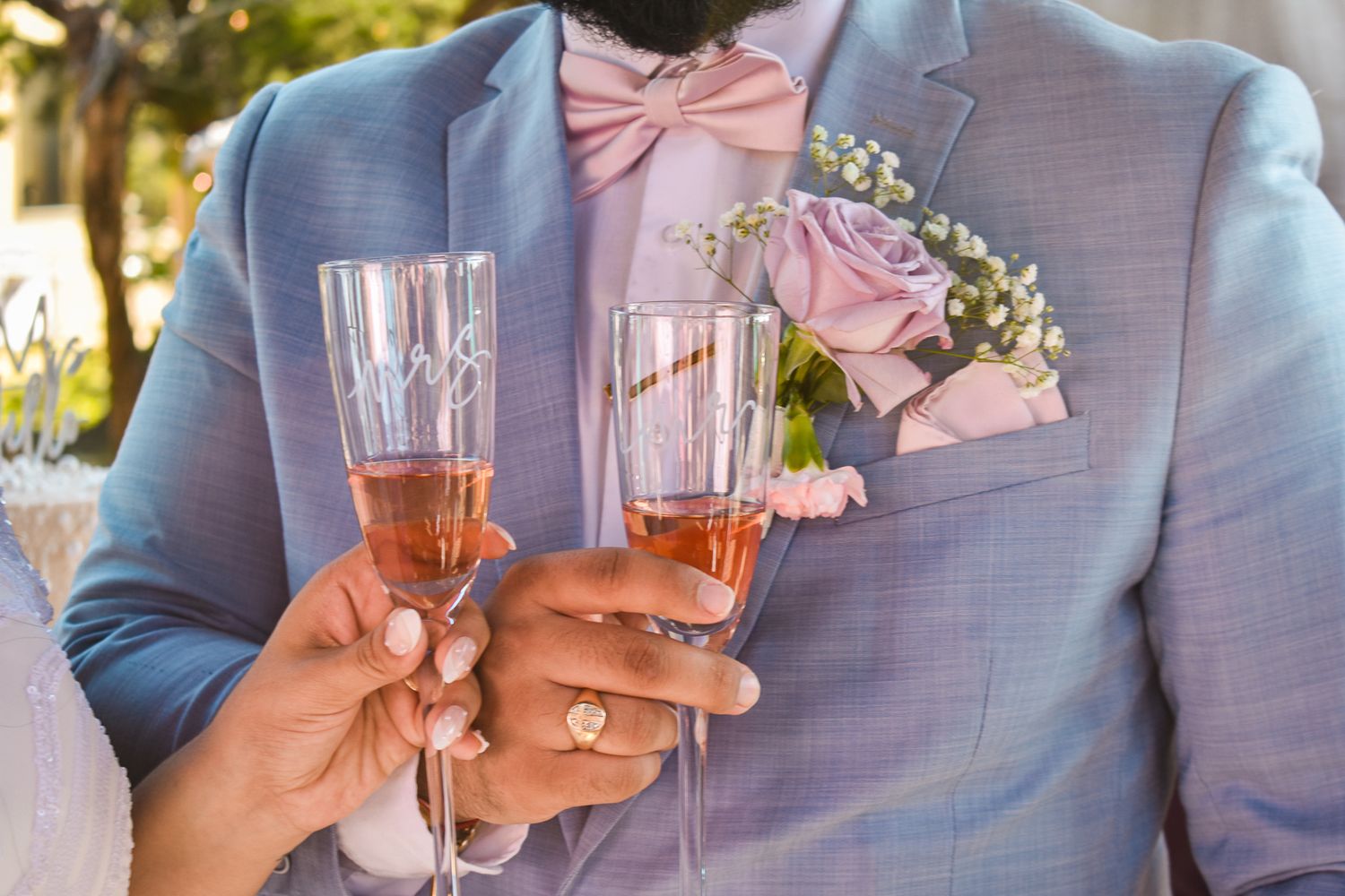 Hands holding champagne flutes with pink boutonniere detail on light blue suit.