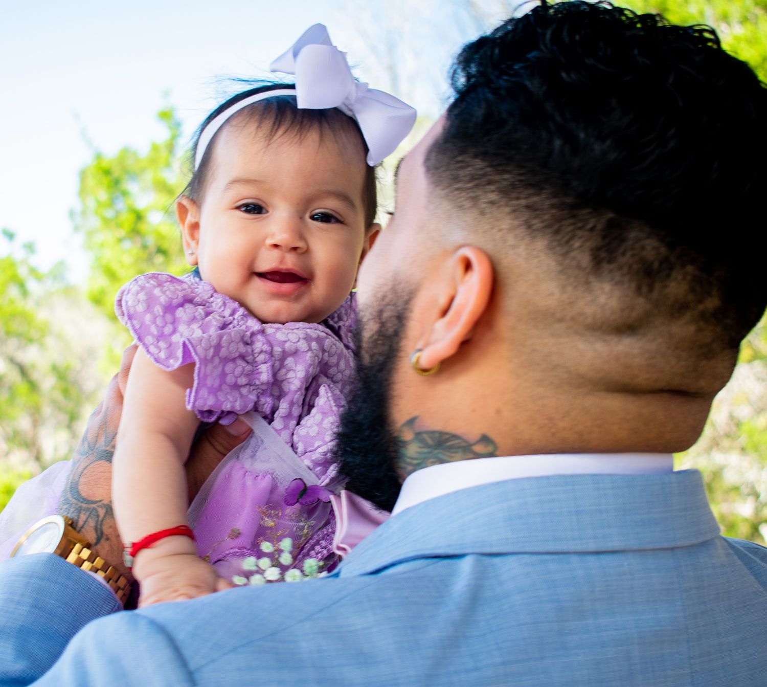 A baby in a purple dress smiles while being held up against a bright outdoor background.