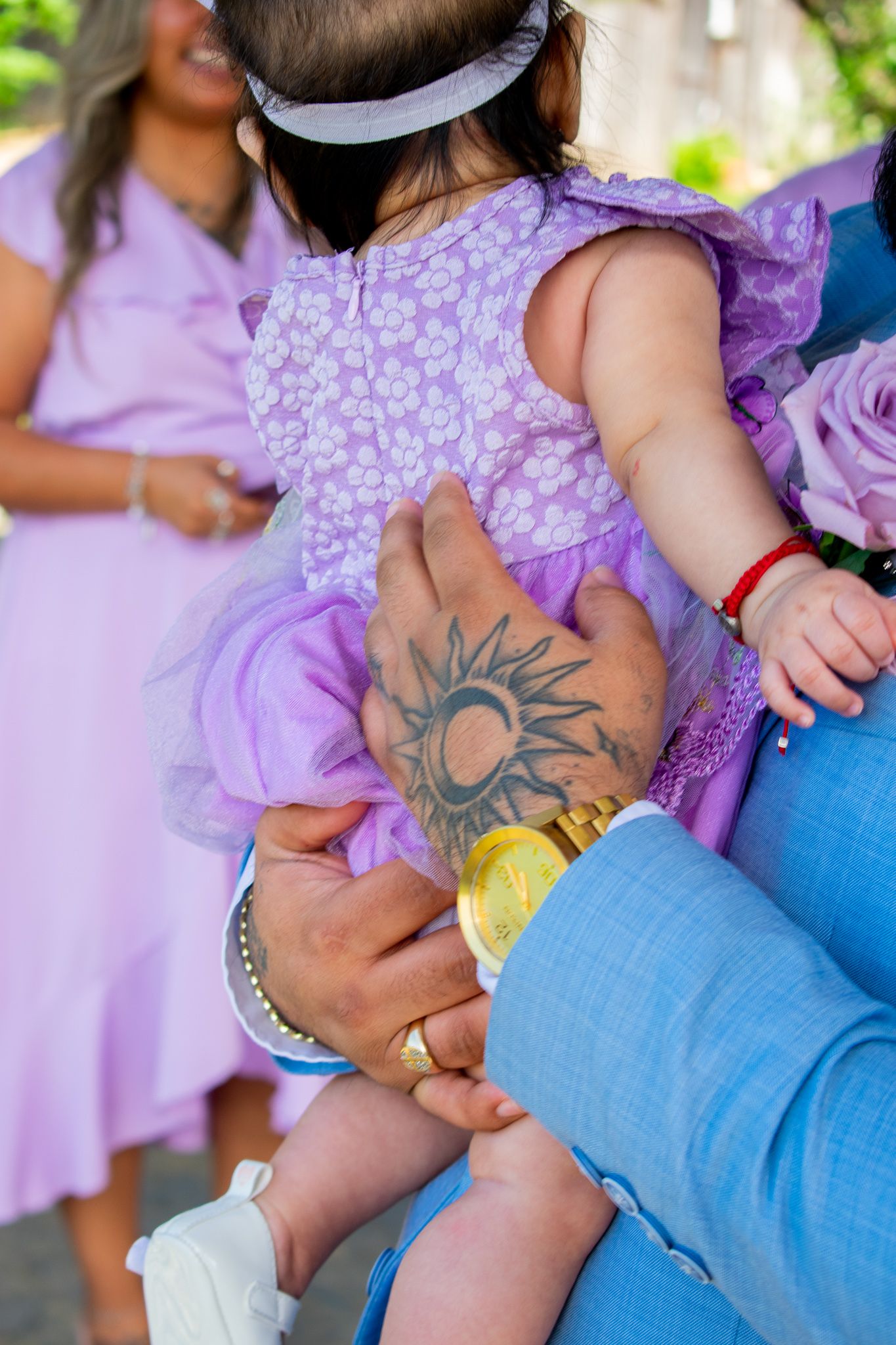 Close-up of hands holding a child in purple dress showing a sun tattoo and jewelry details.