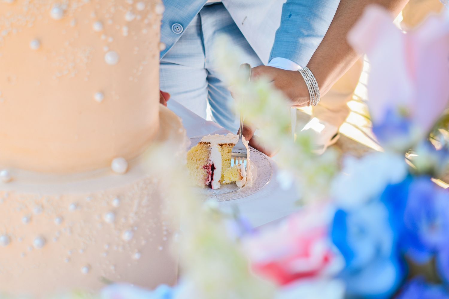 Close-up of hands cutting a white wedding cake surrounded by colorful flowers.