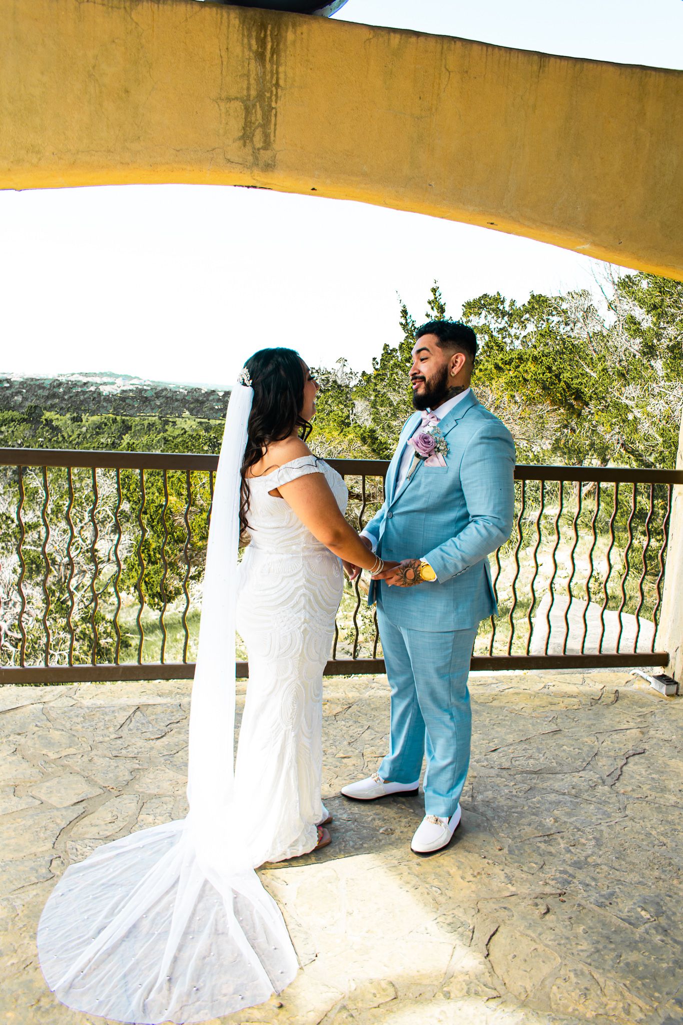 A bride and groom in white dress and light blue suit stand on a balcony overlooking hills.
