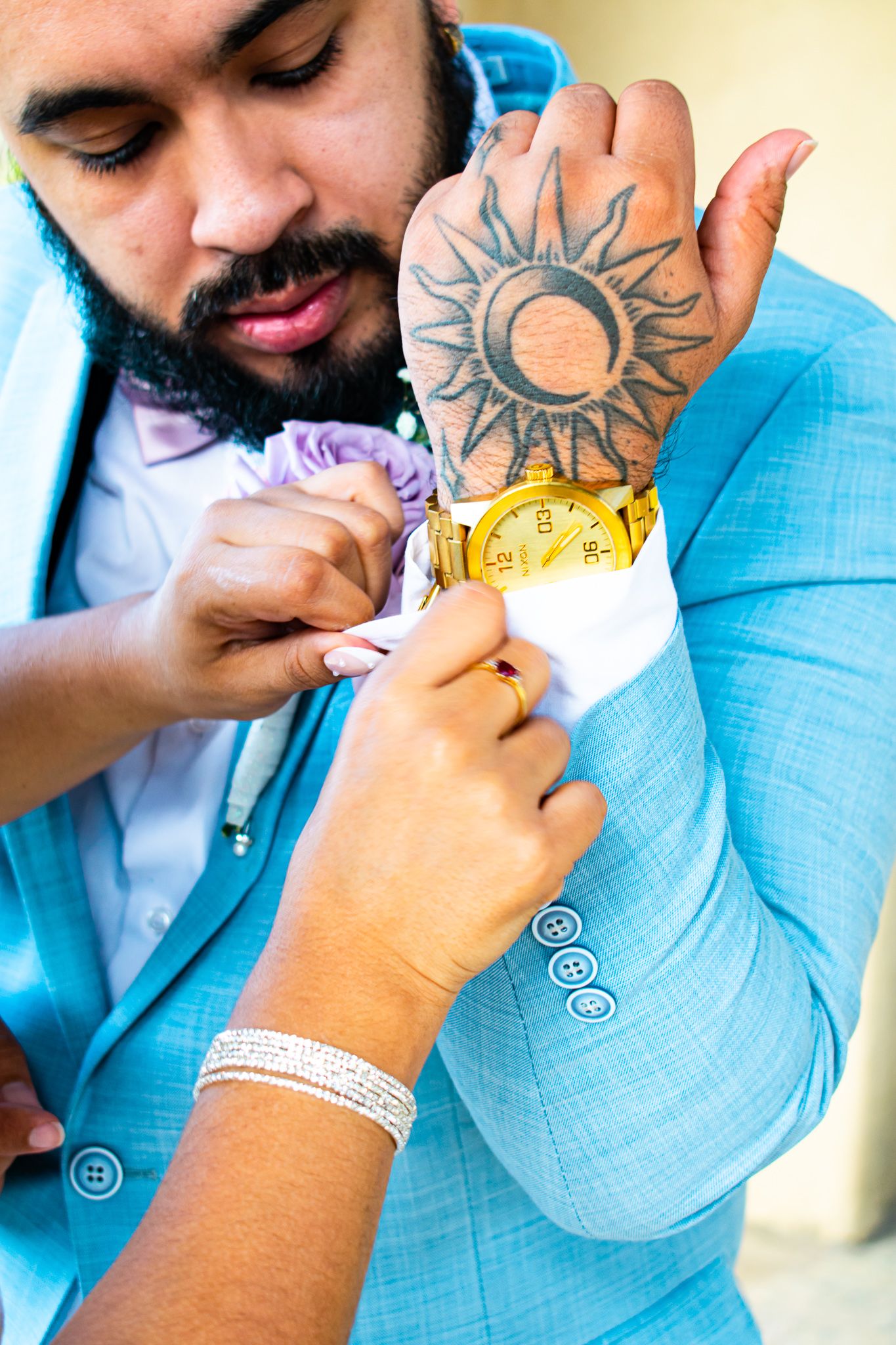 Close-up of hands pinning a boutonniere with a sun tattoo visible on a light blue suit jacket.