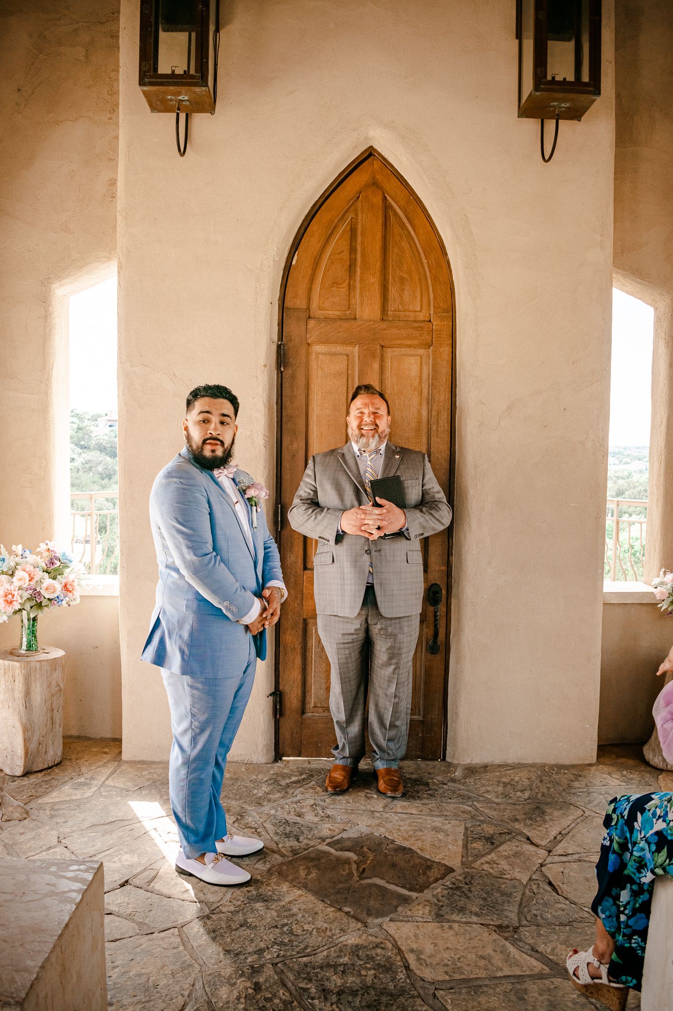 A wedding ceremony setup outside a wooden arched door with wall sconces and natural stone flooring.