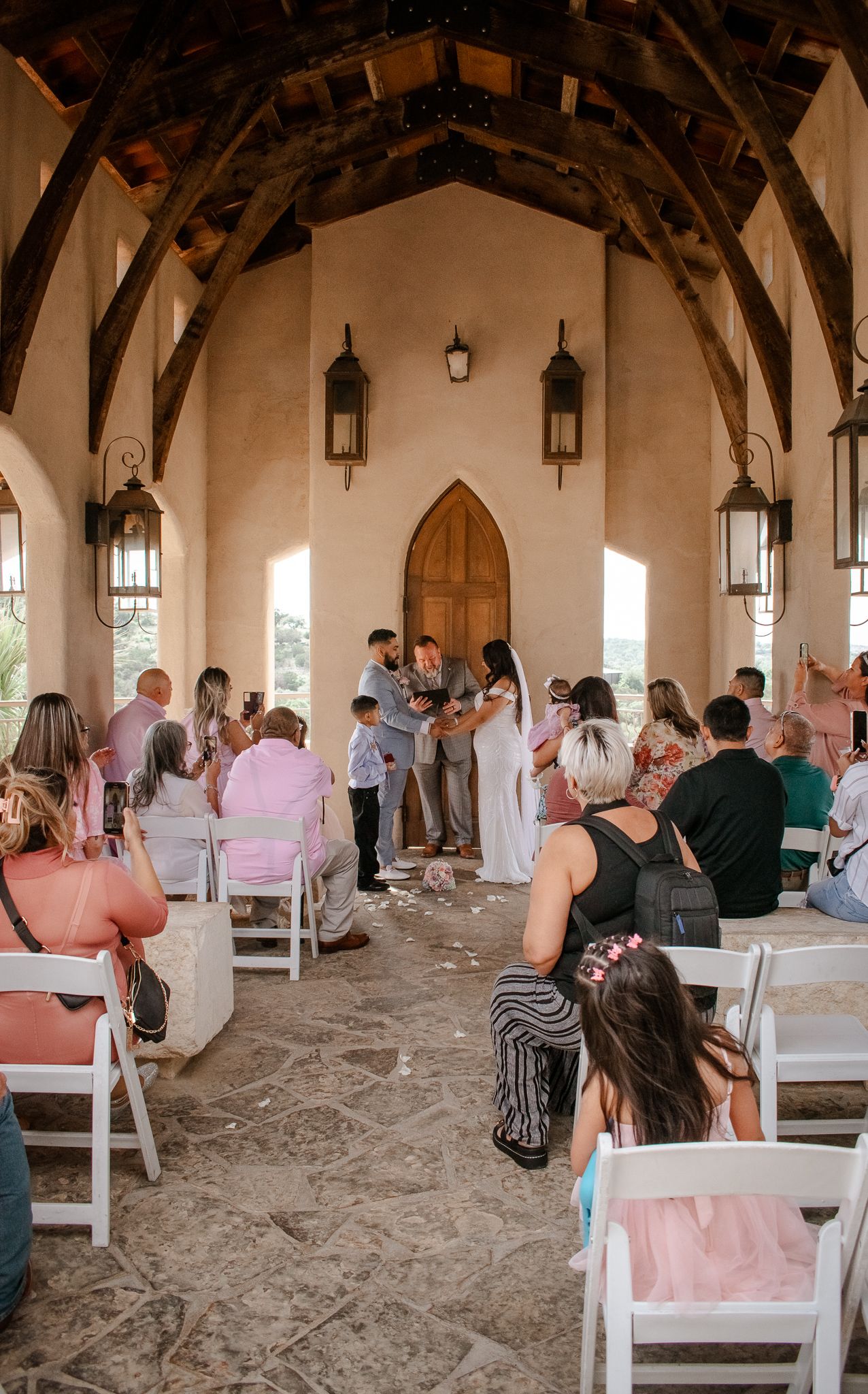 An intimate chapel wedding ceremony with guests seated in white chairs under hanging lanterns.