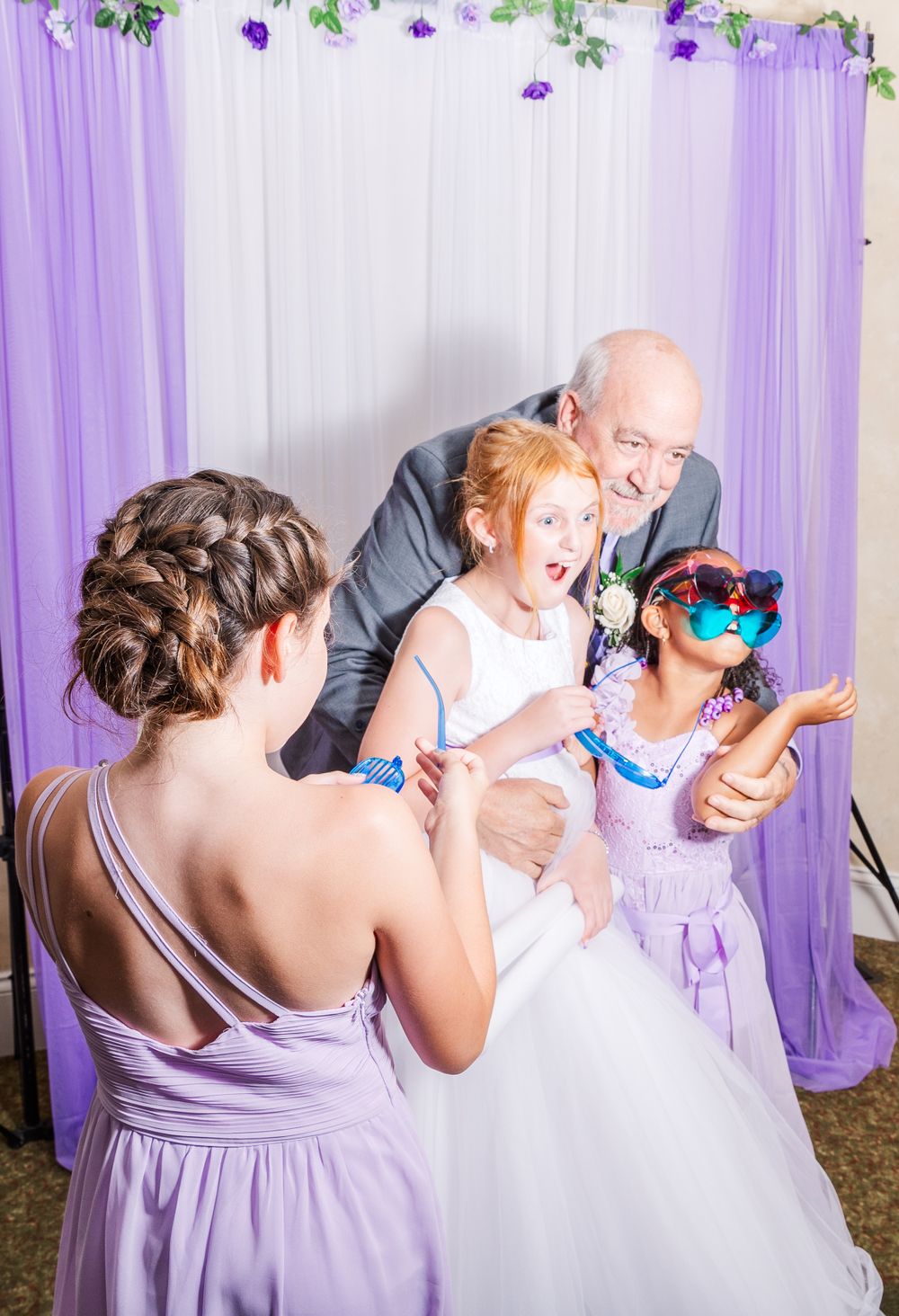 Wedding party poses for photos against a purple and white backdrop with floral decorations.