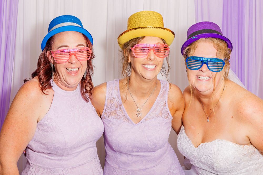 Three friends wearing colorful hats and sunglasses pose together in a wedding photo booth.