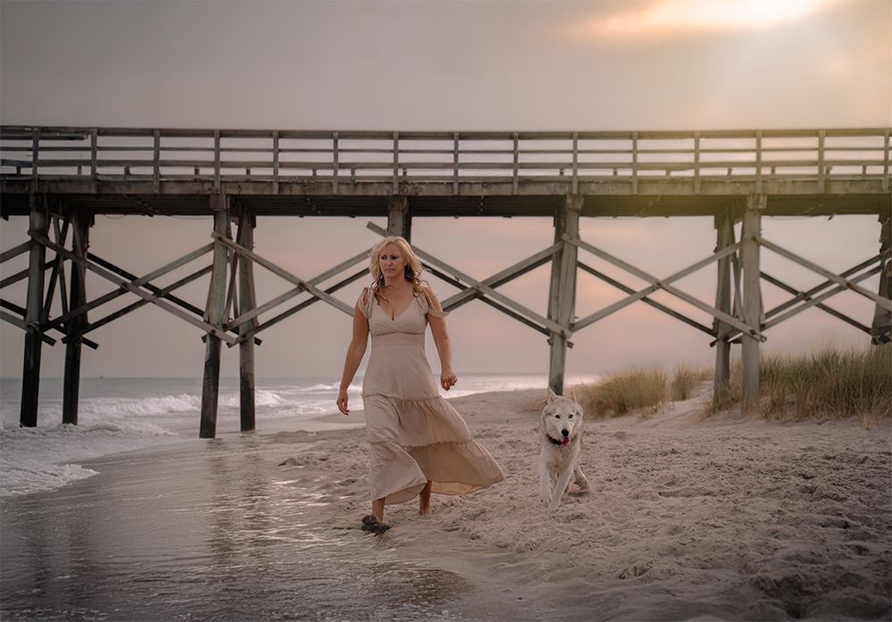 A person in a white dress splashes in shallow water near a wooden pier during sunset.