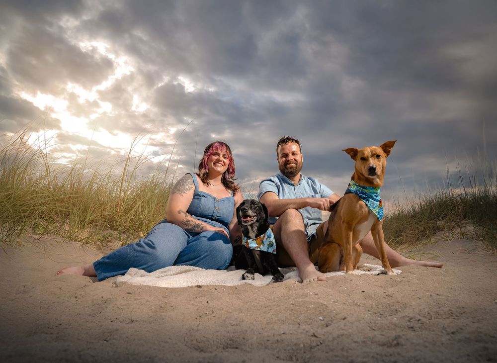 A couple and their dog relax together on a sandy beach at sunset with dramatic cloudy skies overhead.