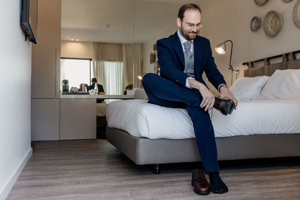Person in navy suit sitting on edge of bed putting on dress shoes.
