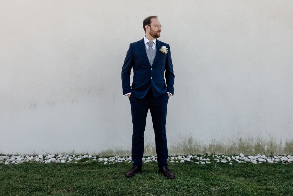 Groom in navy suit standing against neutral wall with landscaping.