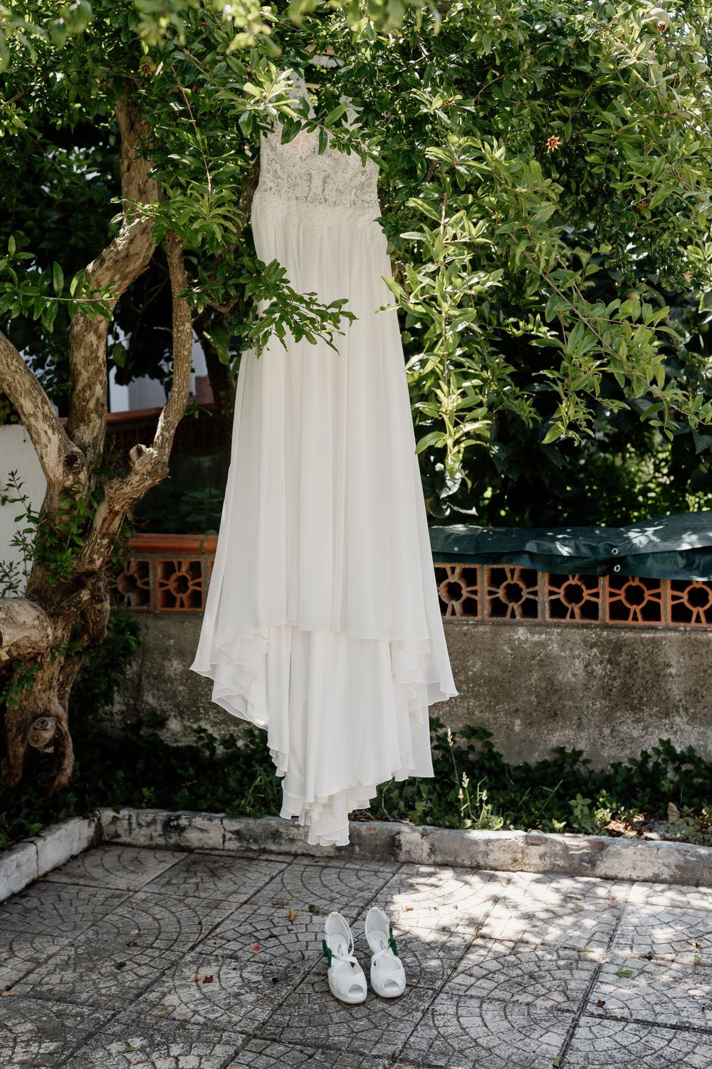 White wedding dress hanging from tree with bridal shoes beneath on stone patio.