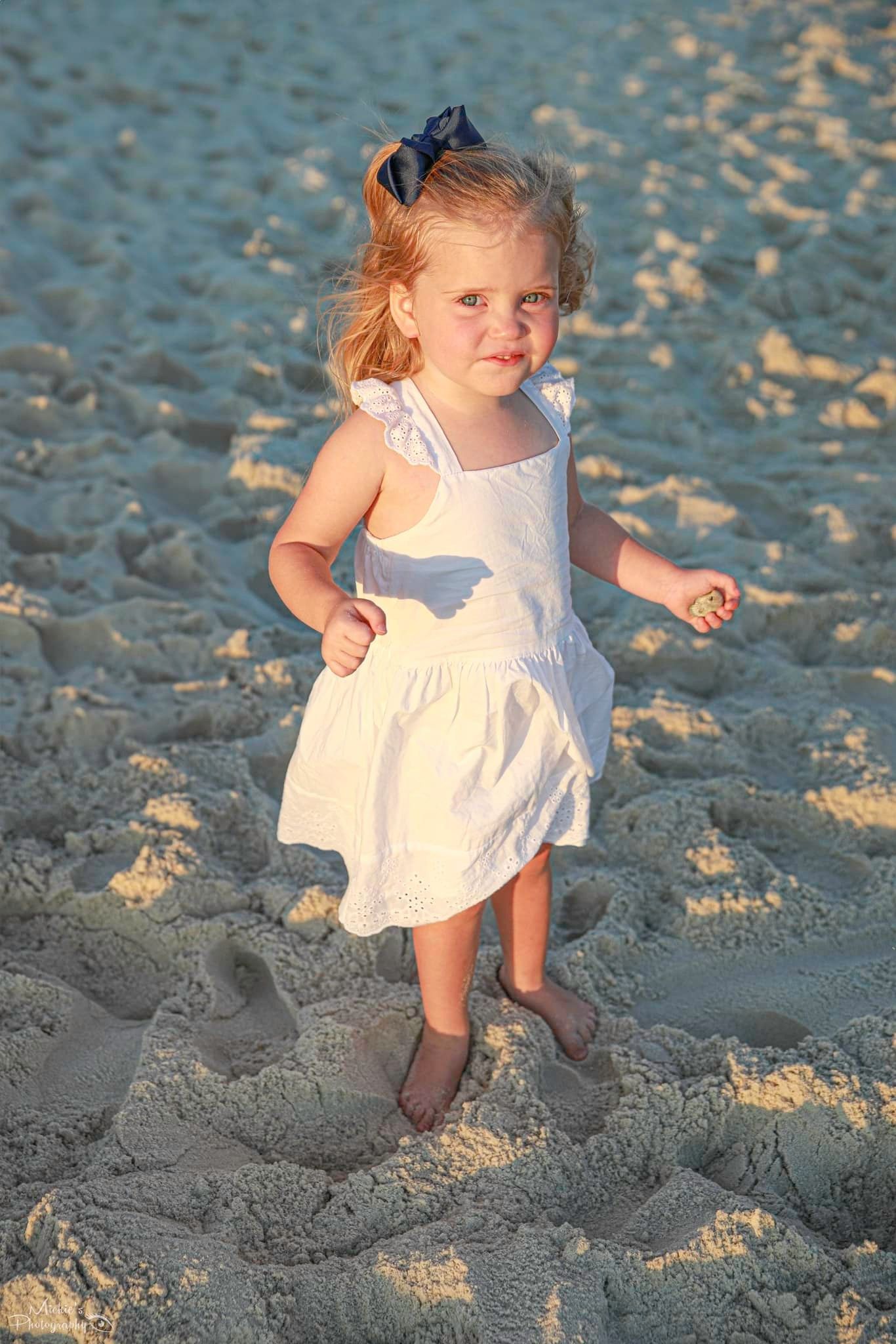 Small child in white dress playing on sandy beach at sunset.