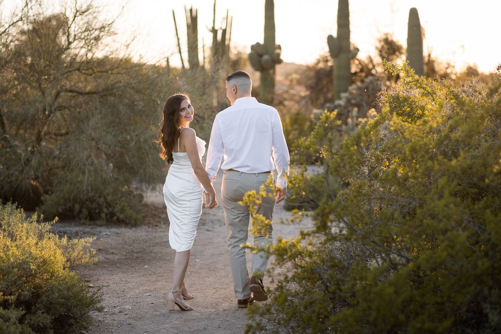 Couple walking hand in hand through desert trail with saguaro cacti at sunset.