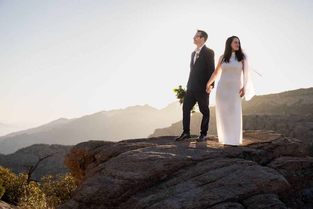 Couple in wedding attire stands on rocky overlook with misty mountains in background.