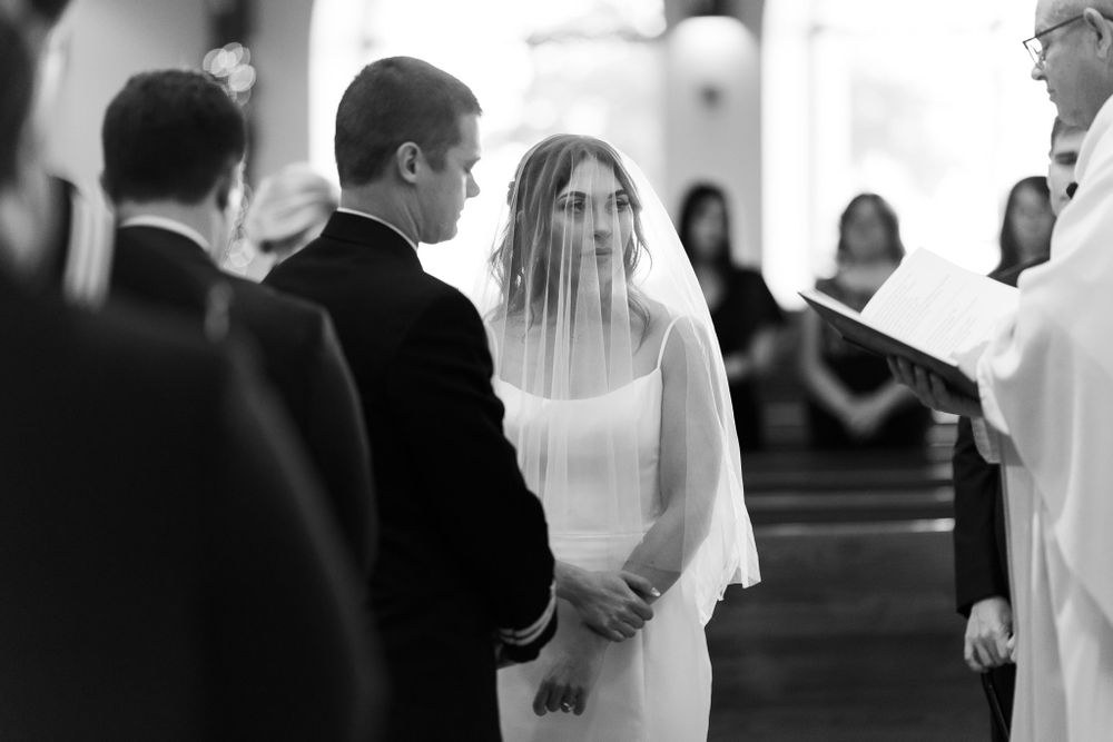 Black and white ceremony photo inside church with traditional wedding attire and priest.