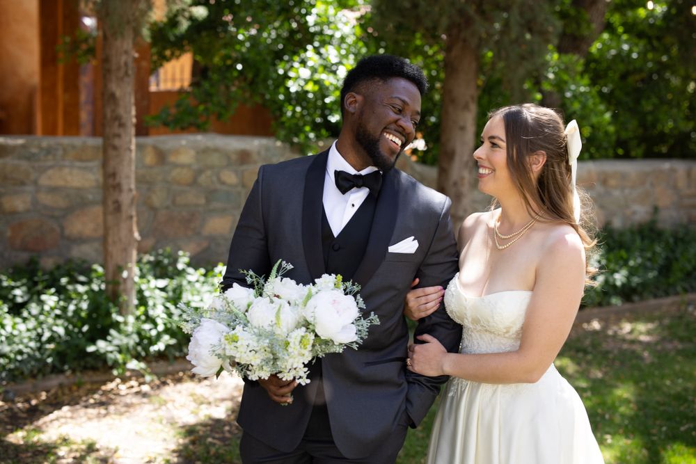 A joyful wedding couple shares a candid laugh in a garden setting with a white floral bouquet.