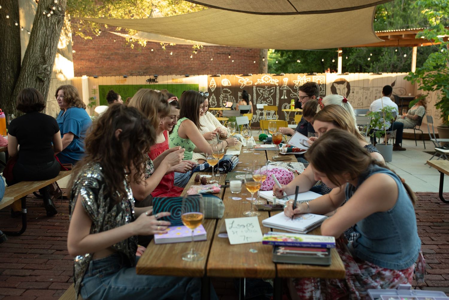 People gather at outdoor tables for a community event under string lights and tree canopy.
