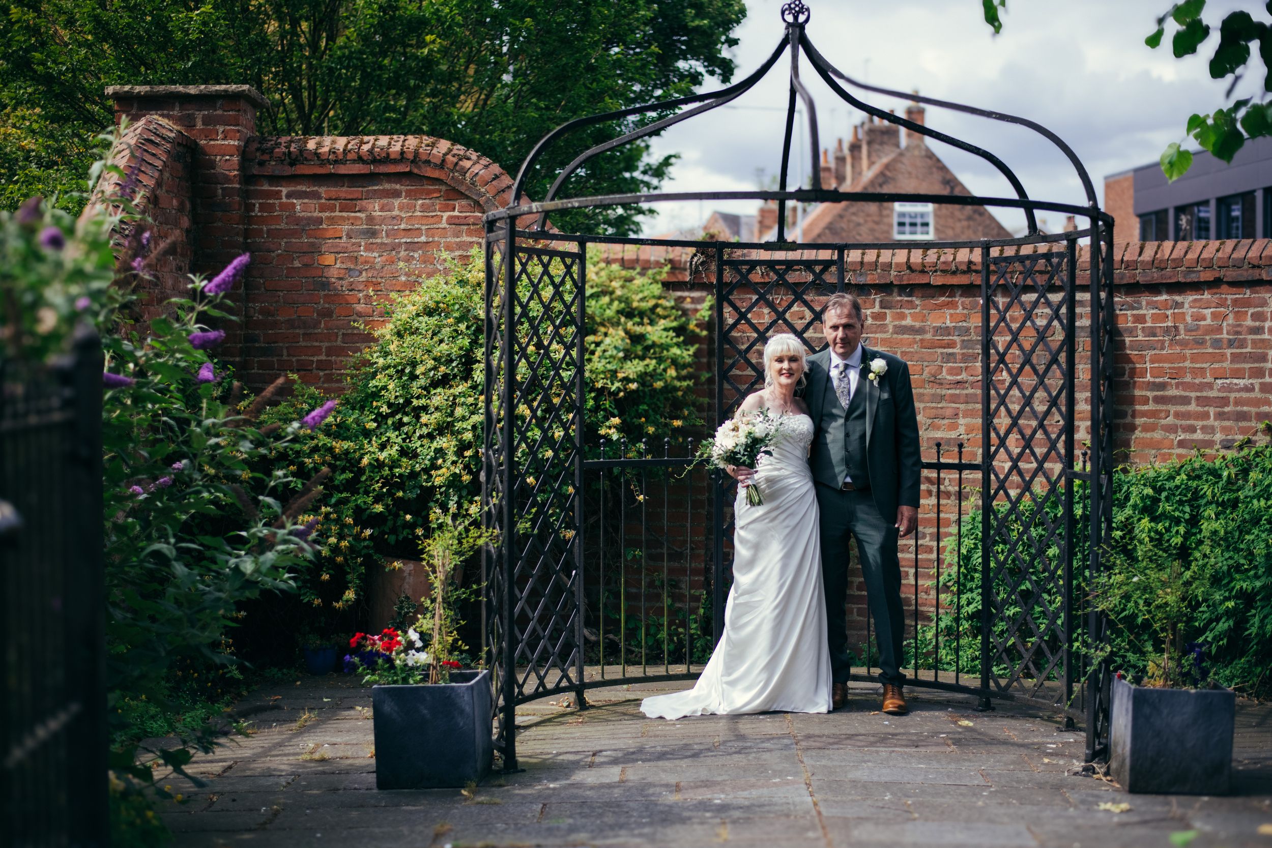 A couple standing under an ornate metal garden archway with brick walls.