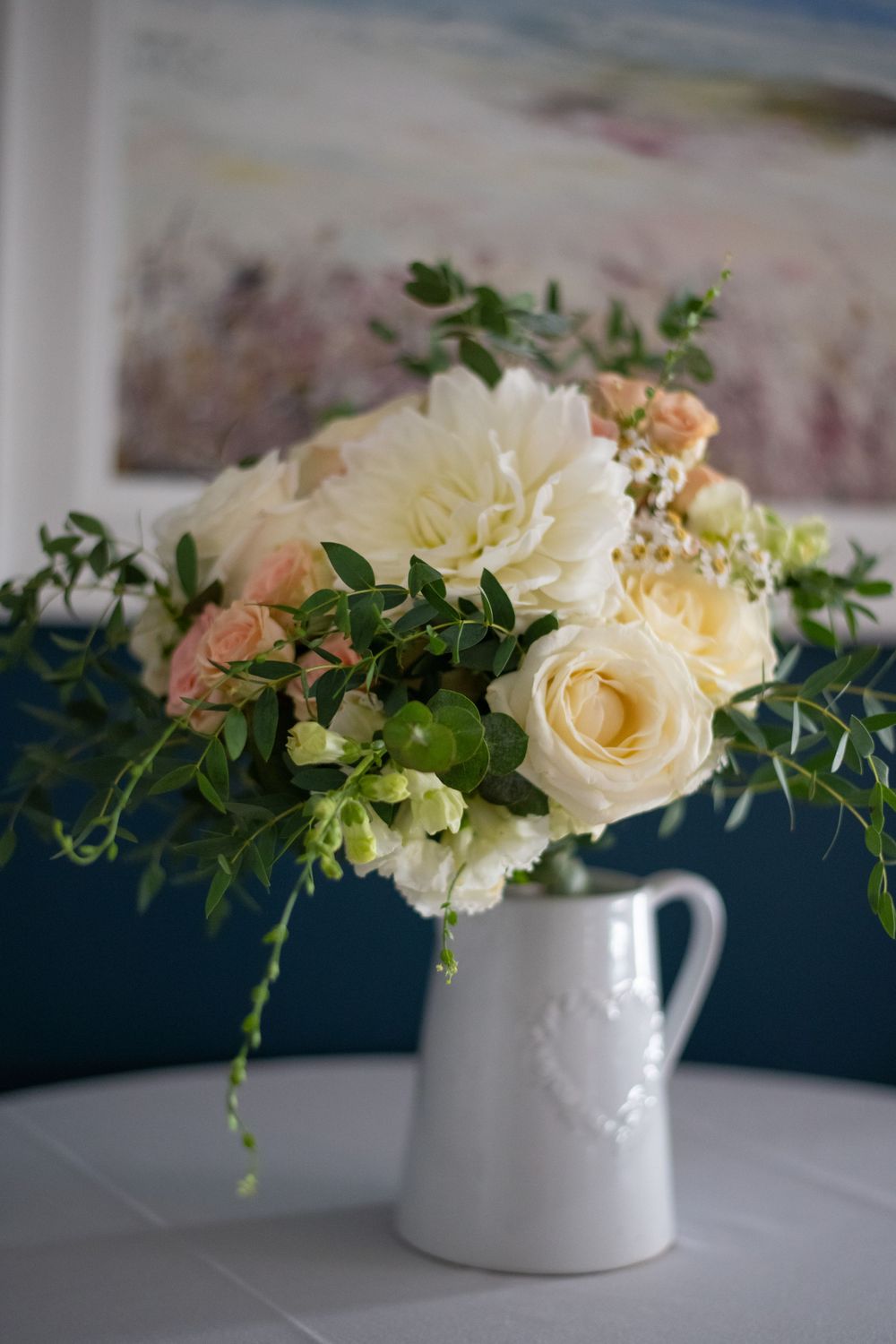 A delicate bouquet of cream roses and greenery arranged in a white ceramic pitcher vase.