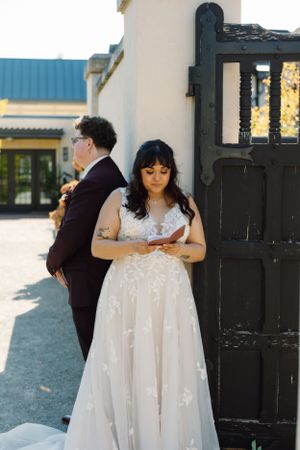 Bride in flowing white lace dress reading their vows near a black gate.