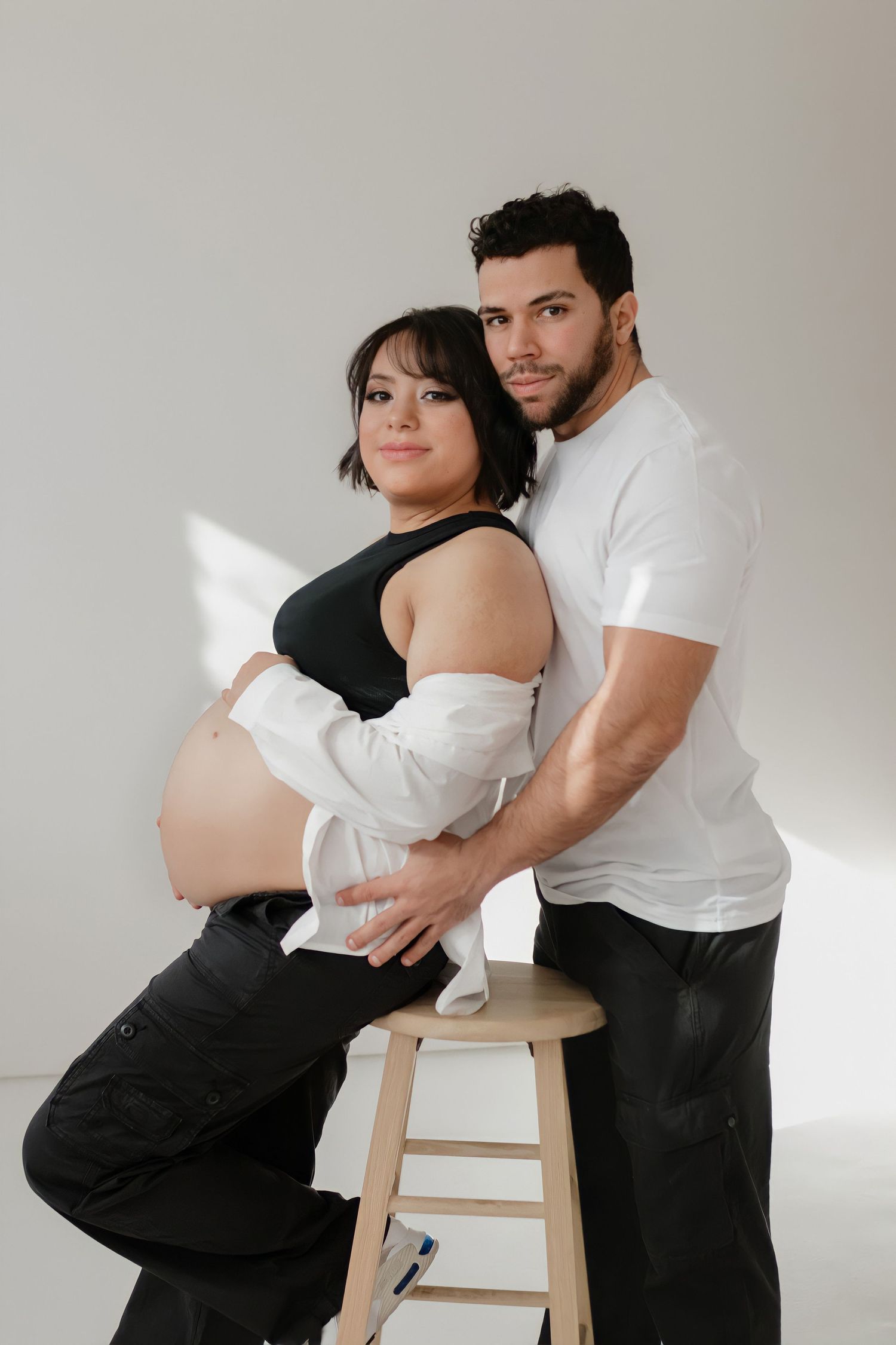 Expectant couple poses in a minimalist studio setting, with her in a black top and white cardigan leaning on a wooden stool.