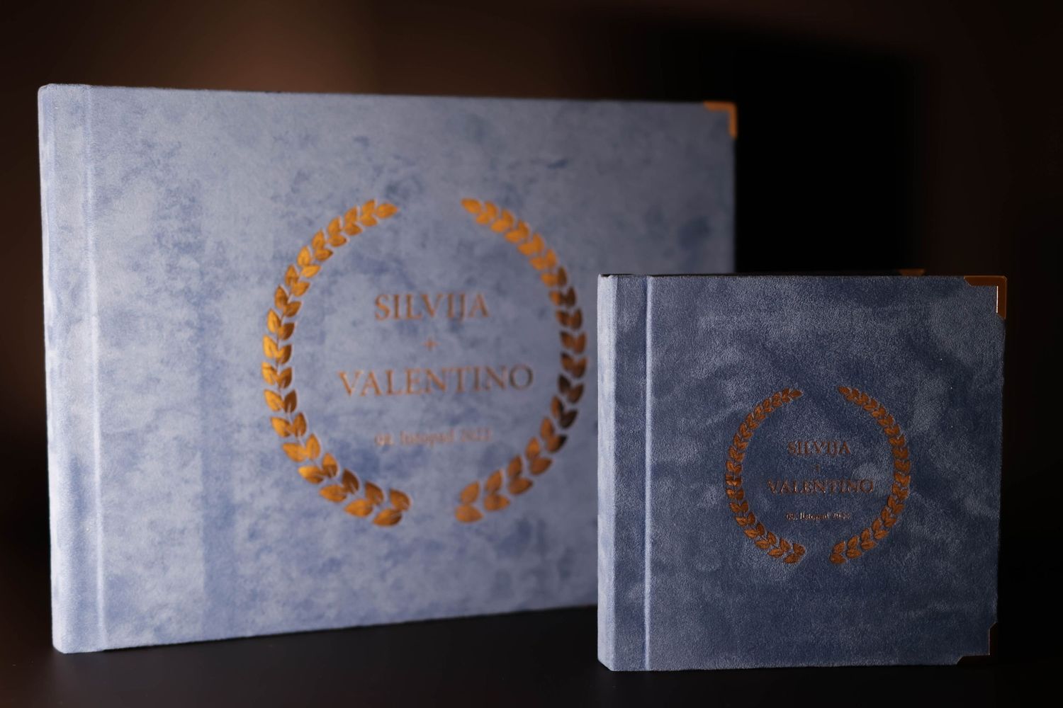 Two elegant blue wedding albums with gold laurel wreath designs on their covers.