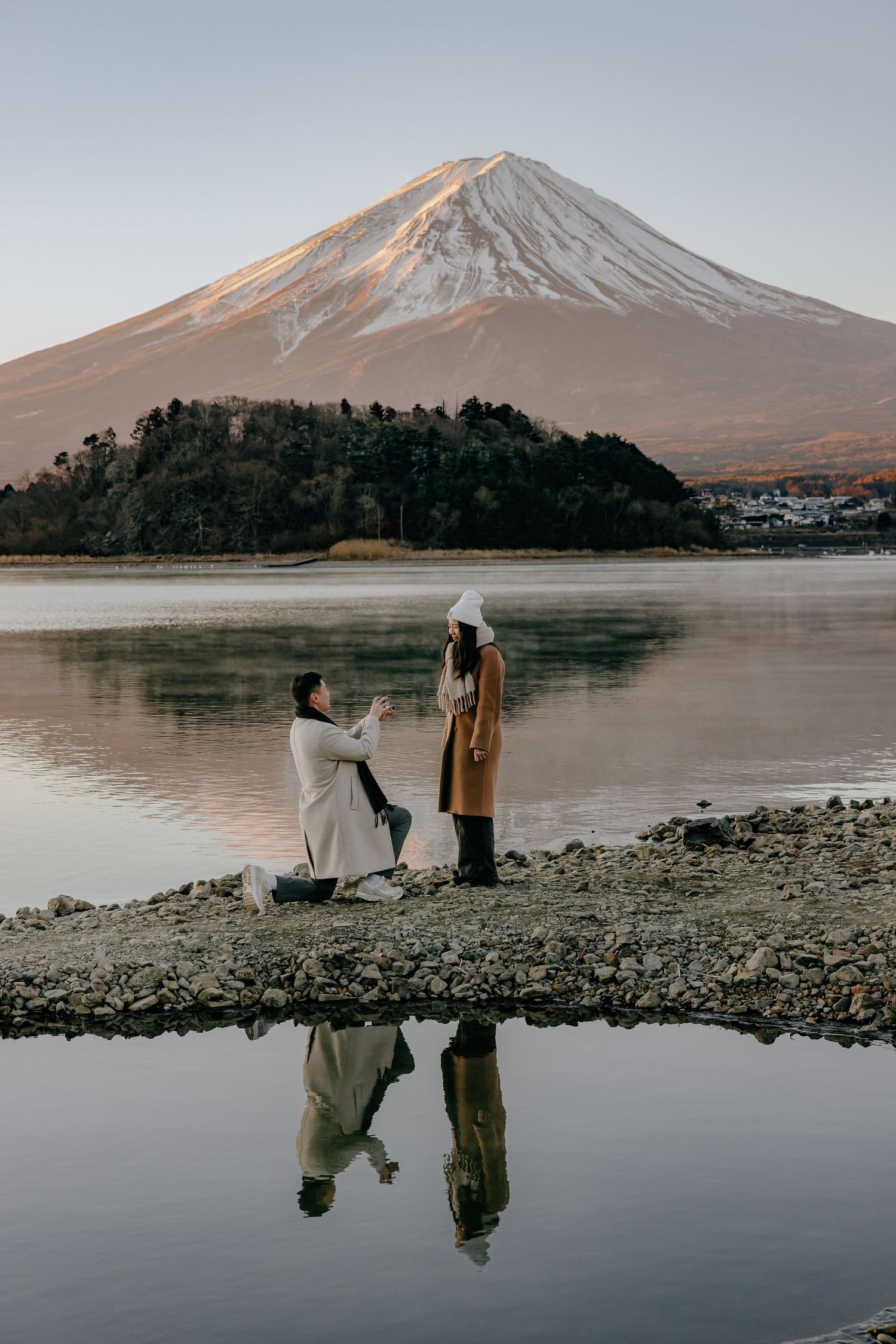 A romantic proposal takes place on the rocky shores of Lake Kawaguchiko with snow-capped Mount Fuji in the background.