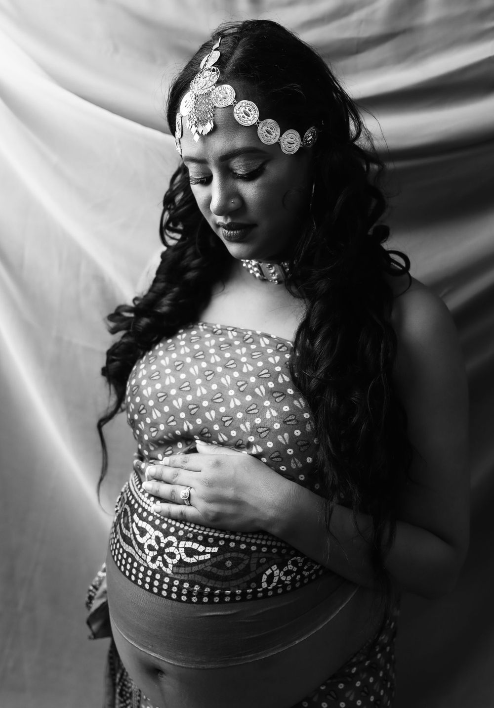 Artistic black and white maternity portrait with ornate head jewelry and embellished outfit.